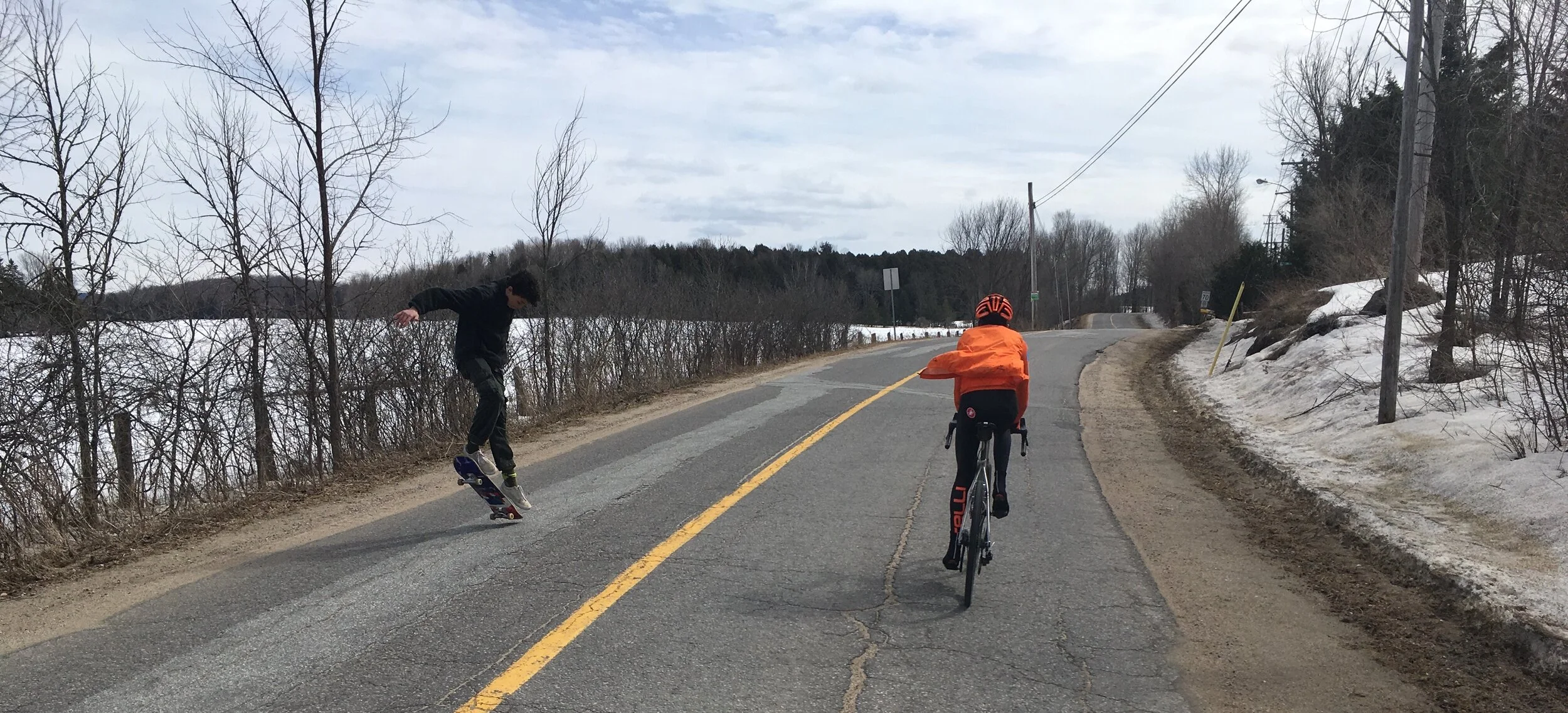 Two children playing on a road with leafless trees and snow on the sides, one riding a bicycle and the other jumping with a skateboard.