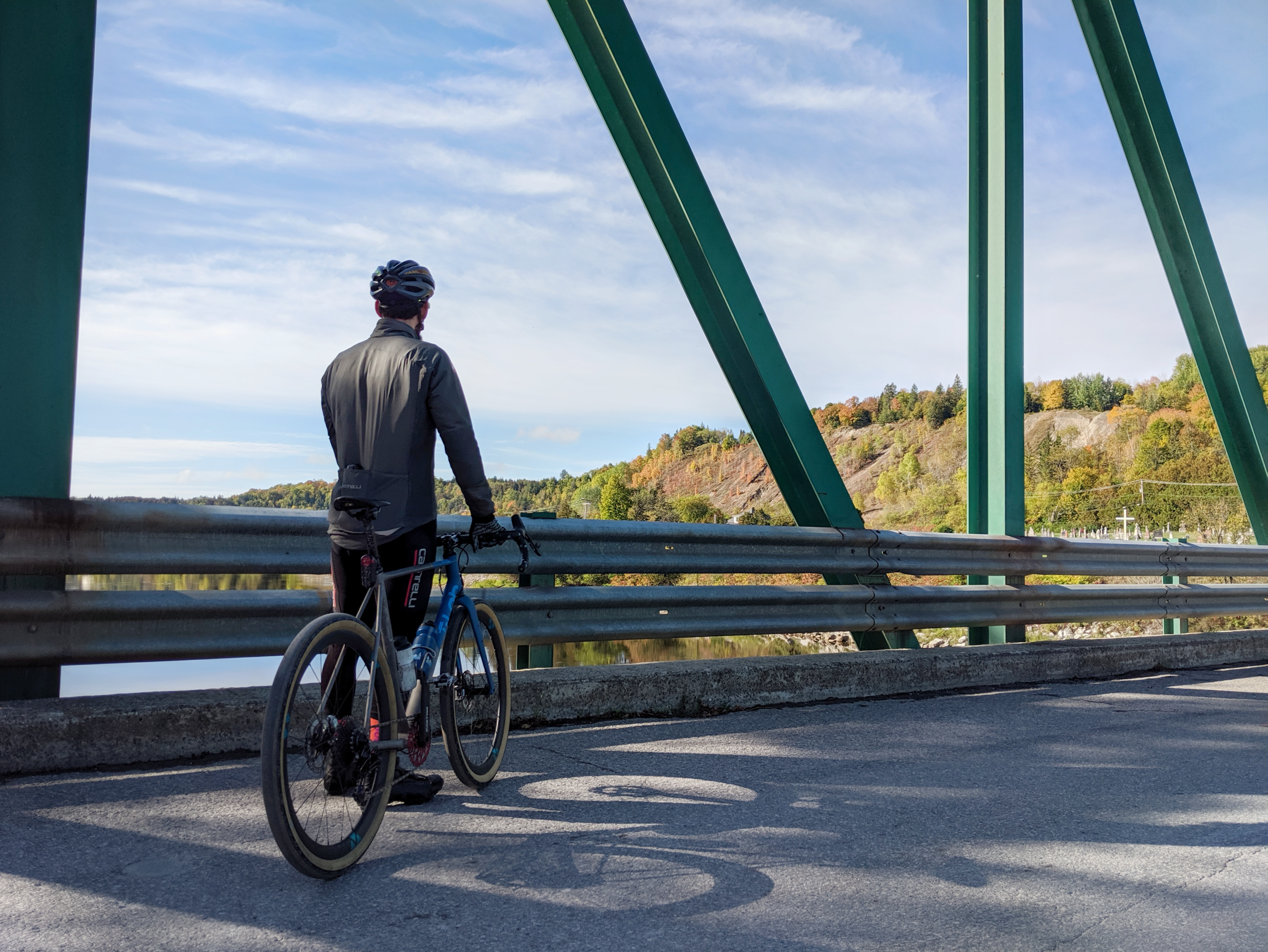 A man wearing a helmet and outdoor jacket stands with his bicycle on a bridge, looking at a scenic view of trees and hills during daytime.