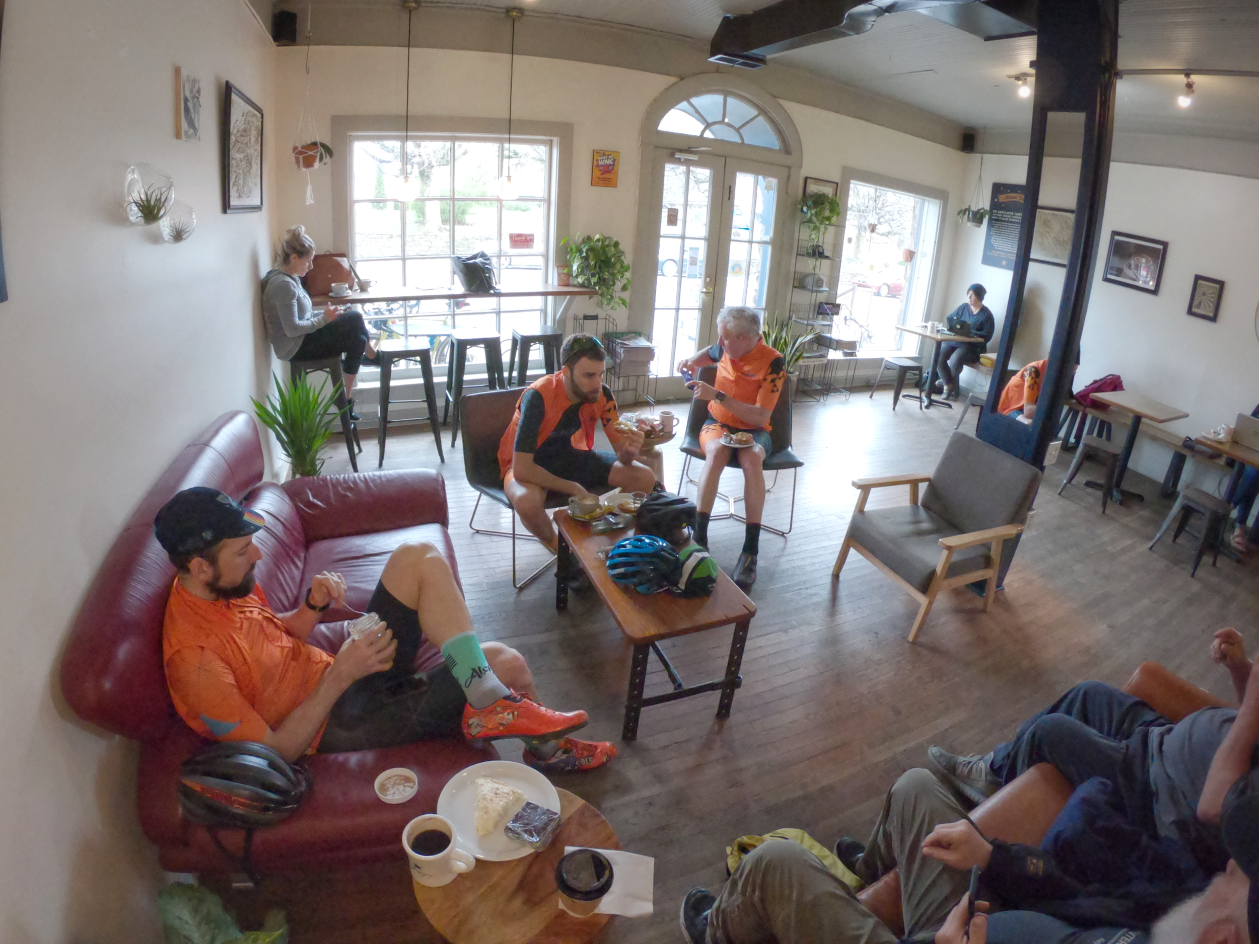 Interior of a cozy café with several people, some wearing colorful cycling gear, sitting and relaxing. A man on a red couch wearing an orange cycling jersey and cap is drinking coffee. Two men in matching orange outfits are seated nearby, one eating 
