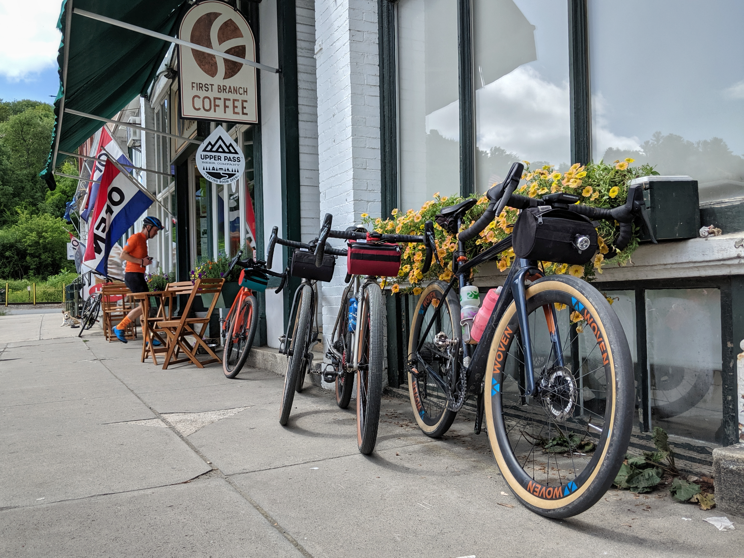 Three bicycles parked outside a coffee shop with a person in cycling gear standing nearby. The shop has a sign that says 'First Branch Coffee' and elements like flags and outdoor seating.