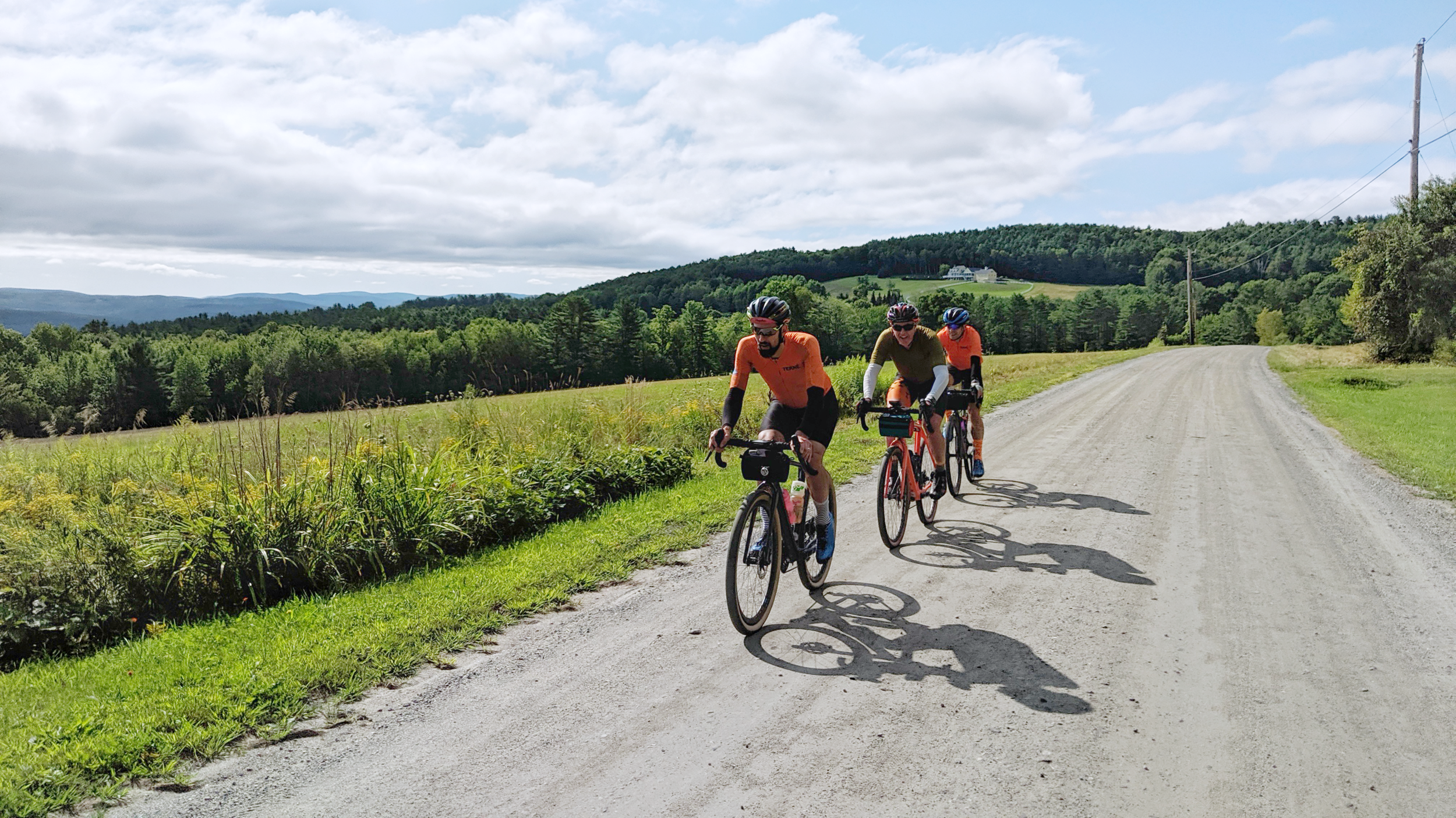 Three cyclists riding on a dirt road through a rural landscape with green fields, trees, and hills in the background under a partly cloudy sky.