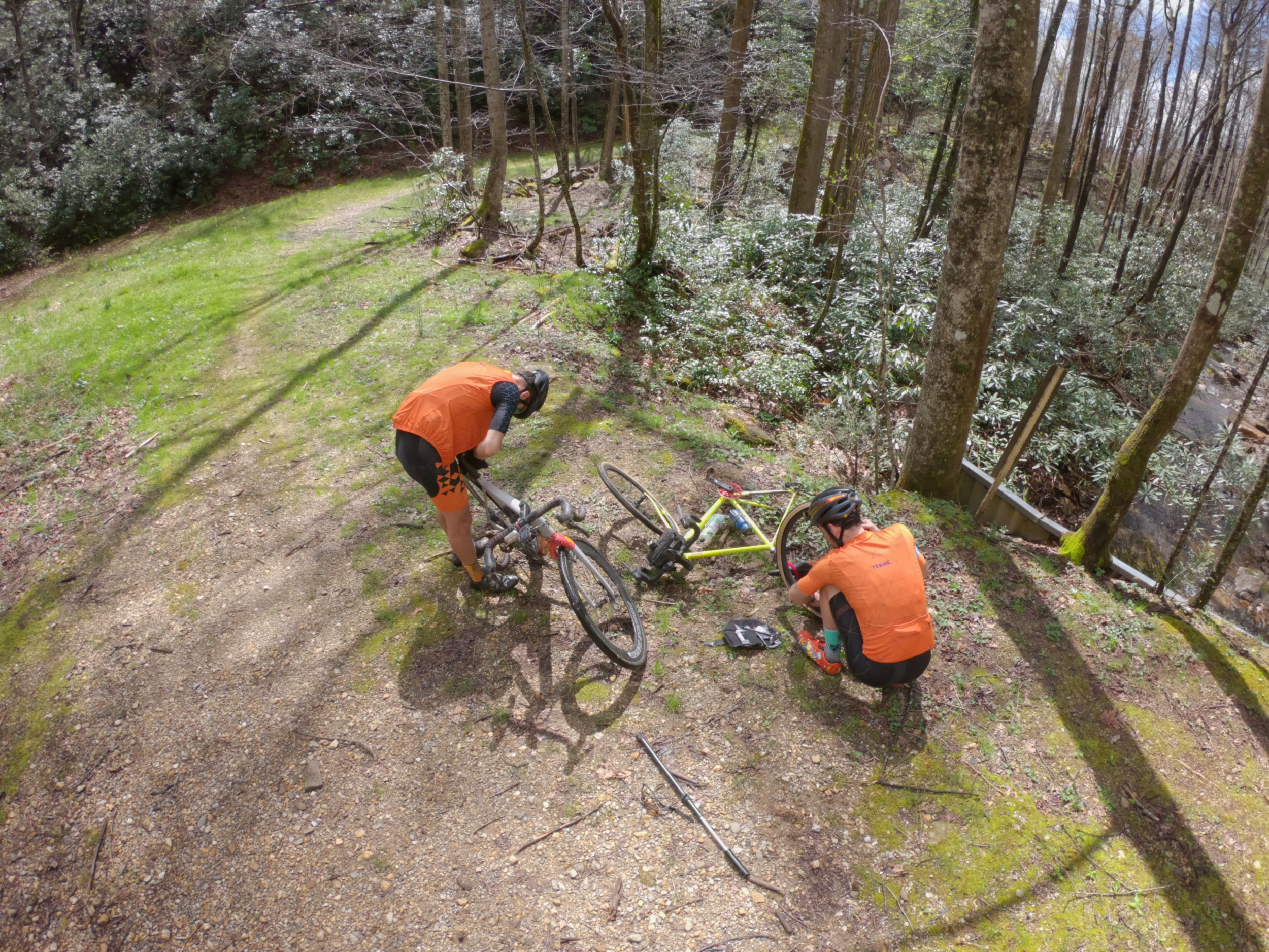 Two mountain bikers repairing a yellow mountain bike on a forest trail, with trees and a creek nearby.