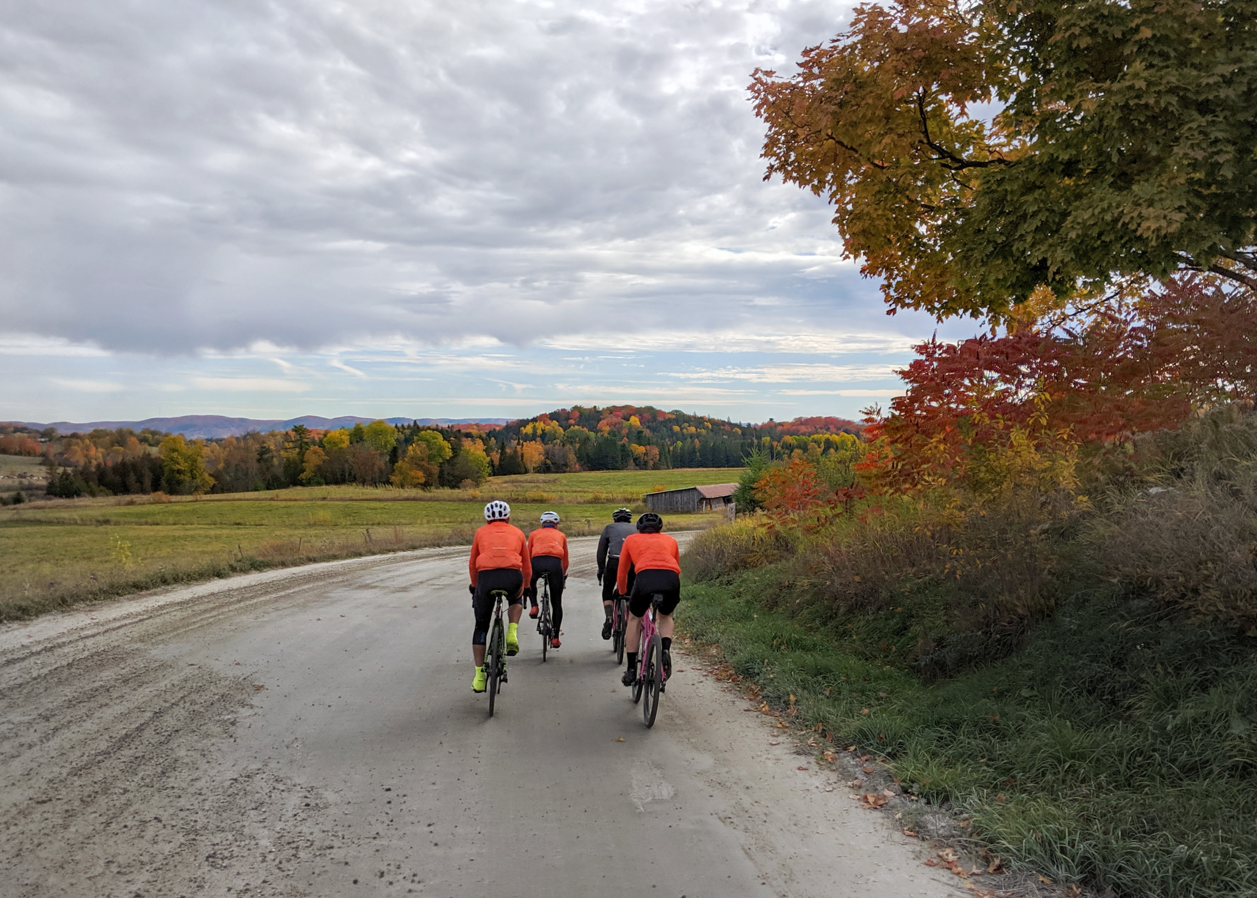 Four bikers riding on a rural dirt road surrounded by fall foliage and rolling hills under a cloudy sky.