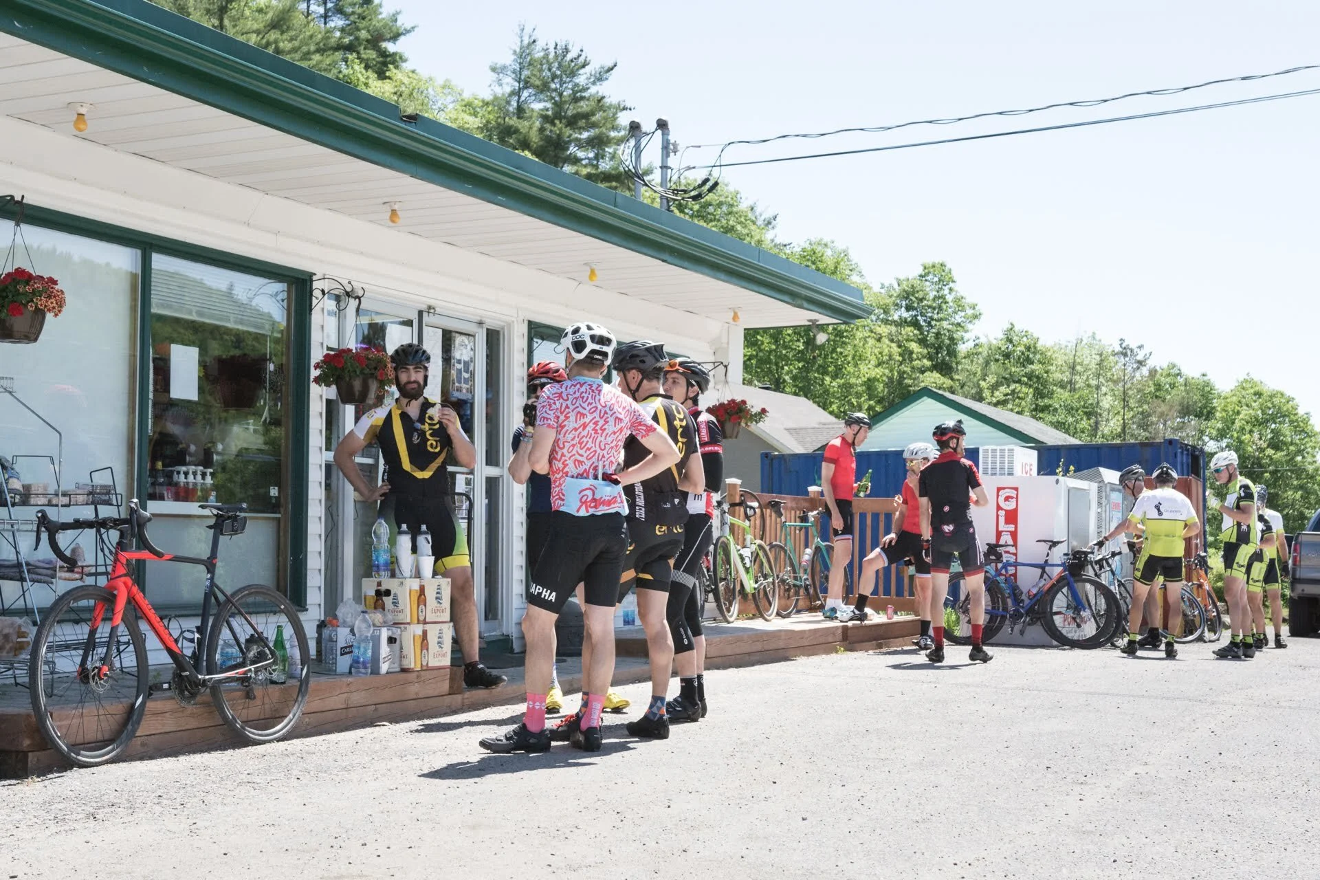 Cyclists gathered outside a small convenience store, some drinking water and chatting, with bicycles parked nearby on a sunny day.