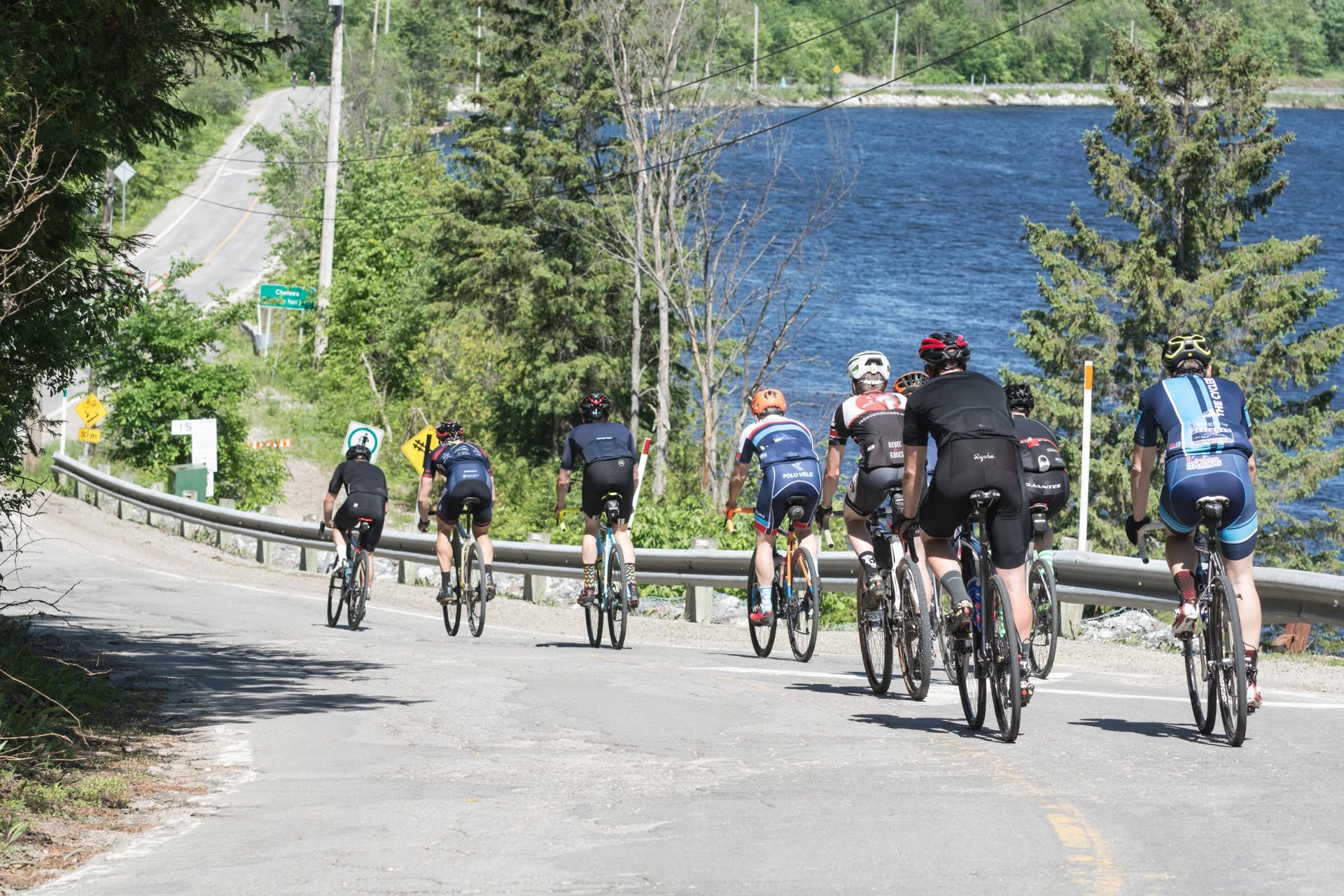A group of cyclists riding up a hill on a paved road near a lake, surrounded by green trees, with road signs and a guardrail along the roadside.