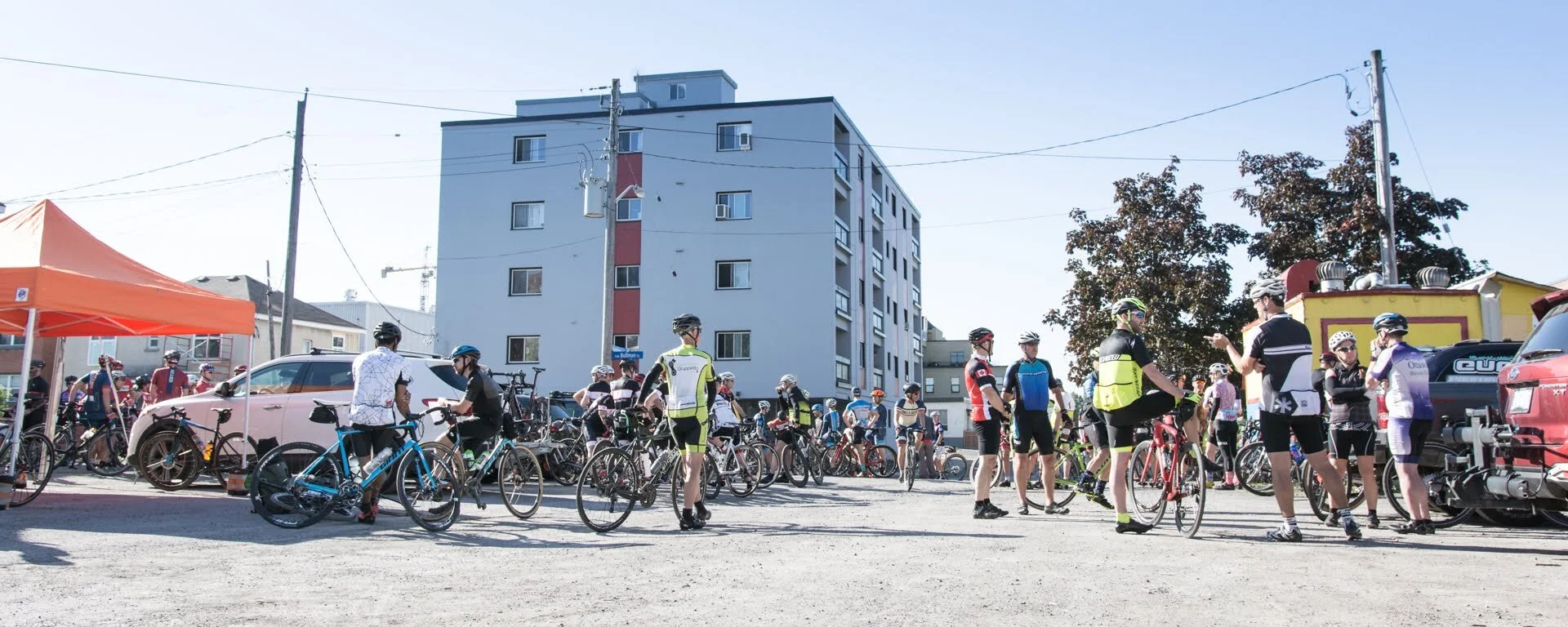 Group of cyclists gathering outside in a parking lot under a clear sky, with multi-story buildings and trees in the background, and a bright orange tent on the left.