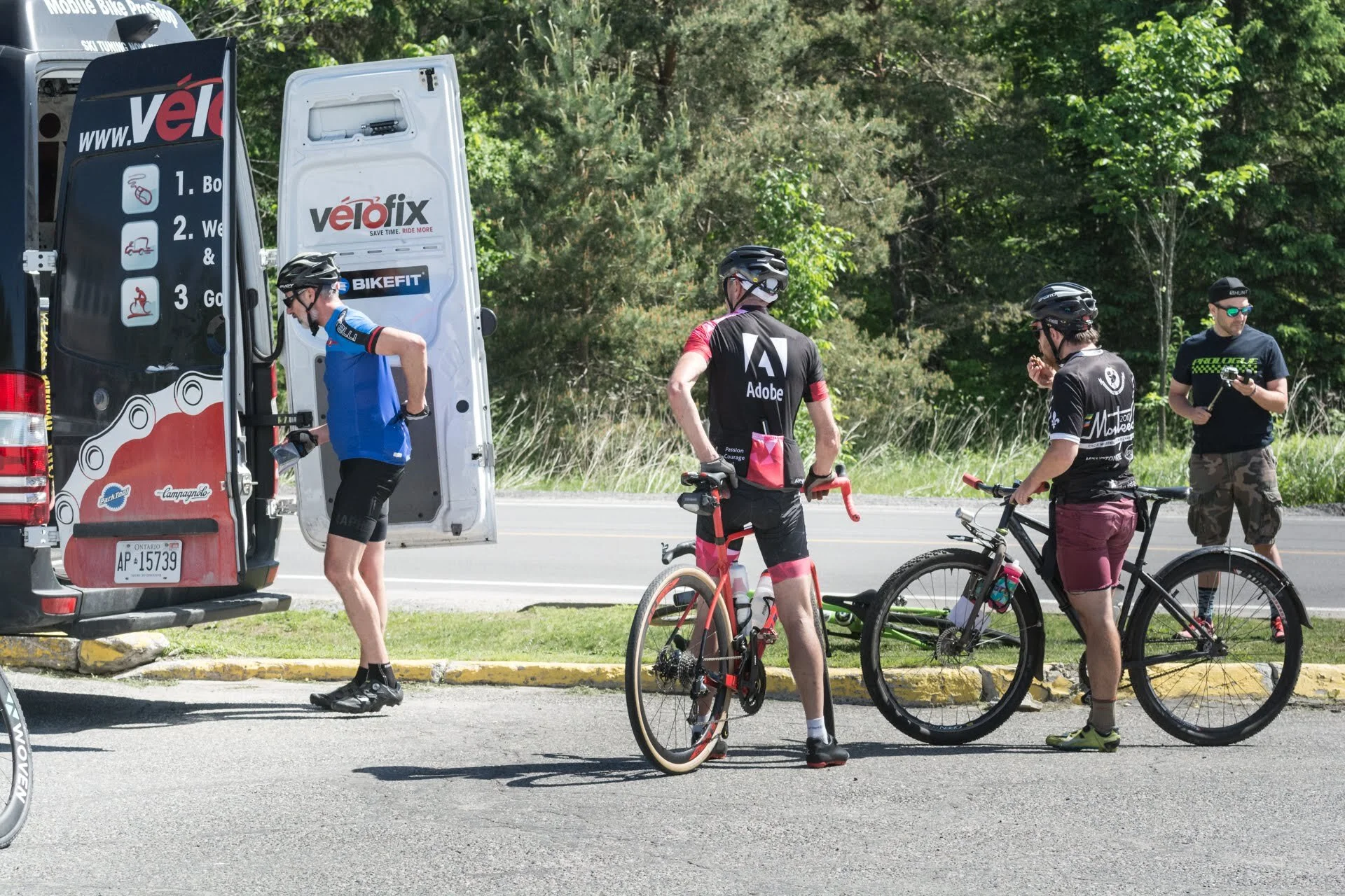 A group of four cyclists in sports gear preparing near a van with an open door, surrounded by greenery on a sunny day.