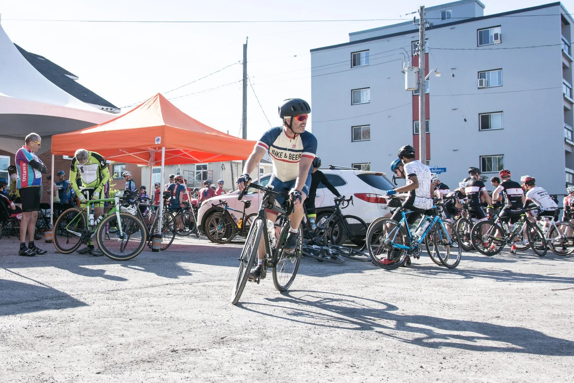 Cyclists gather at a cycling event with a man riding a bike in the foreground. There are multiple bikes and people in cycling gear, with a tent and a building in the background.