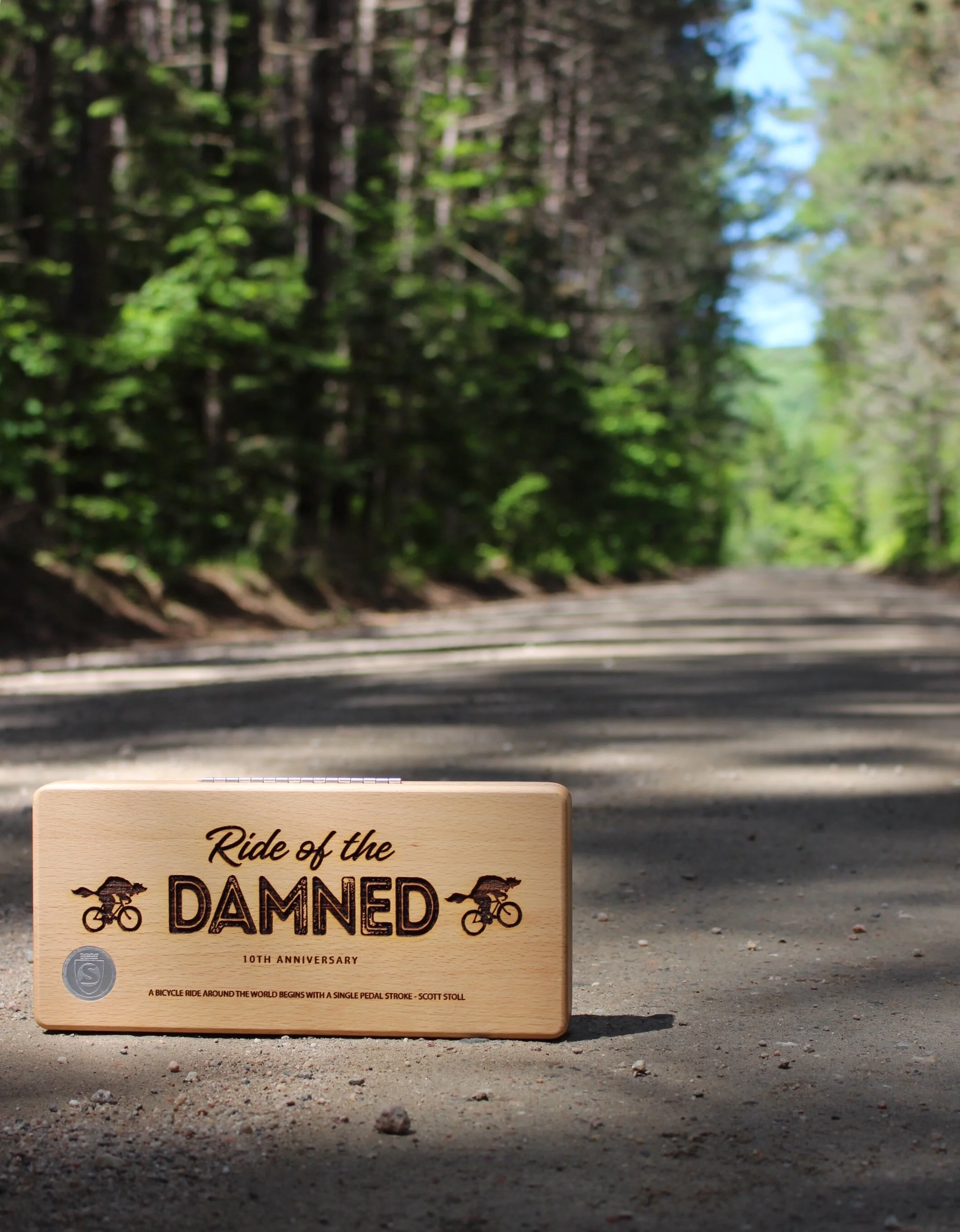 A wooden plaque on the ground in a forested area with trees and a dirt road in the background. The plaque reads: 'Ride of the DAMNED 10th Anniversary' and includes images of bicycles and a quote.
