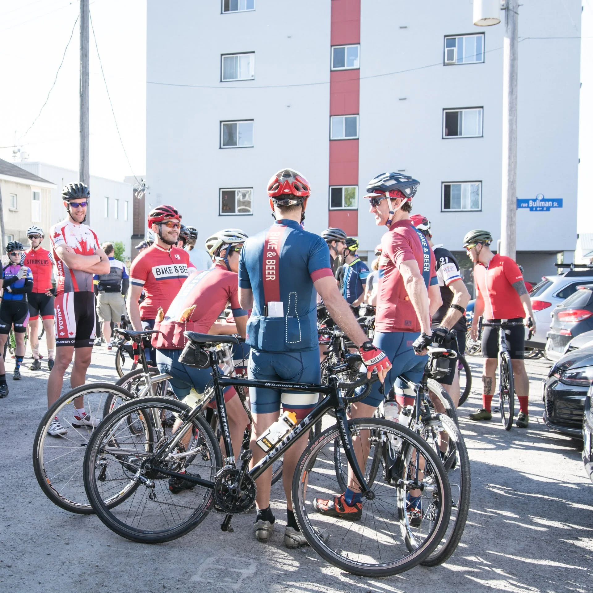 Cyclists gathered in a parking lot, preparing for a ride, many wearing red, blue, and black cycling jerseys and helmets.