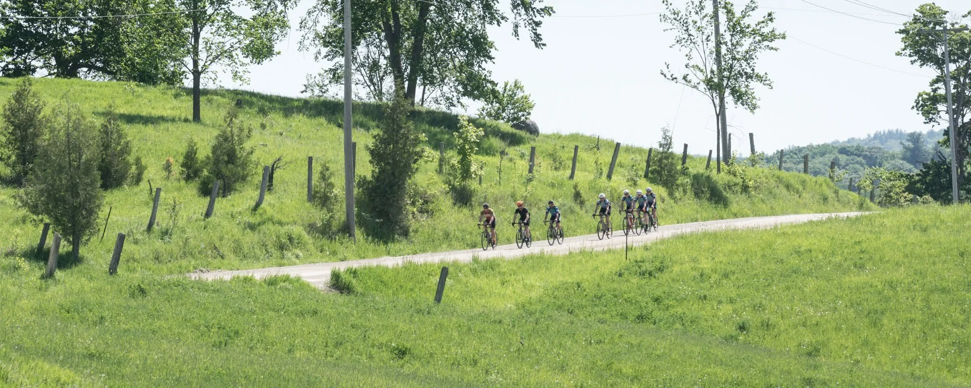 Group of six cyclists riding on a paved road through a green rural landscape with trees and grassy hills.