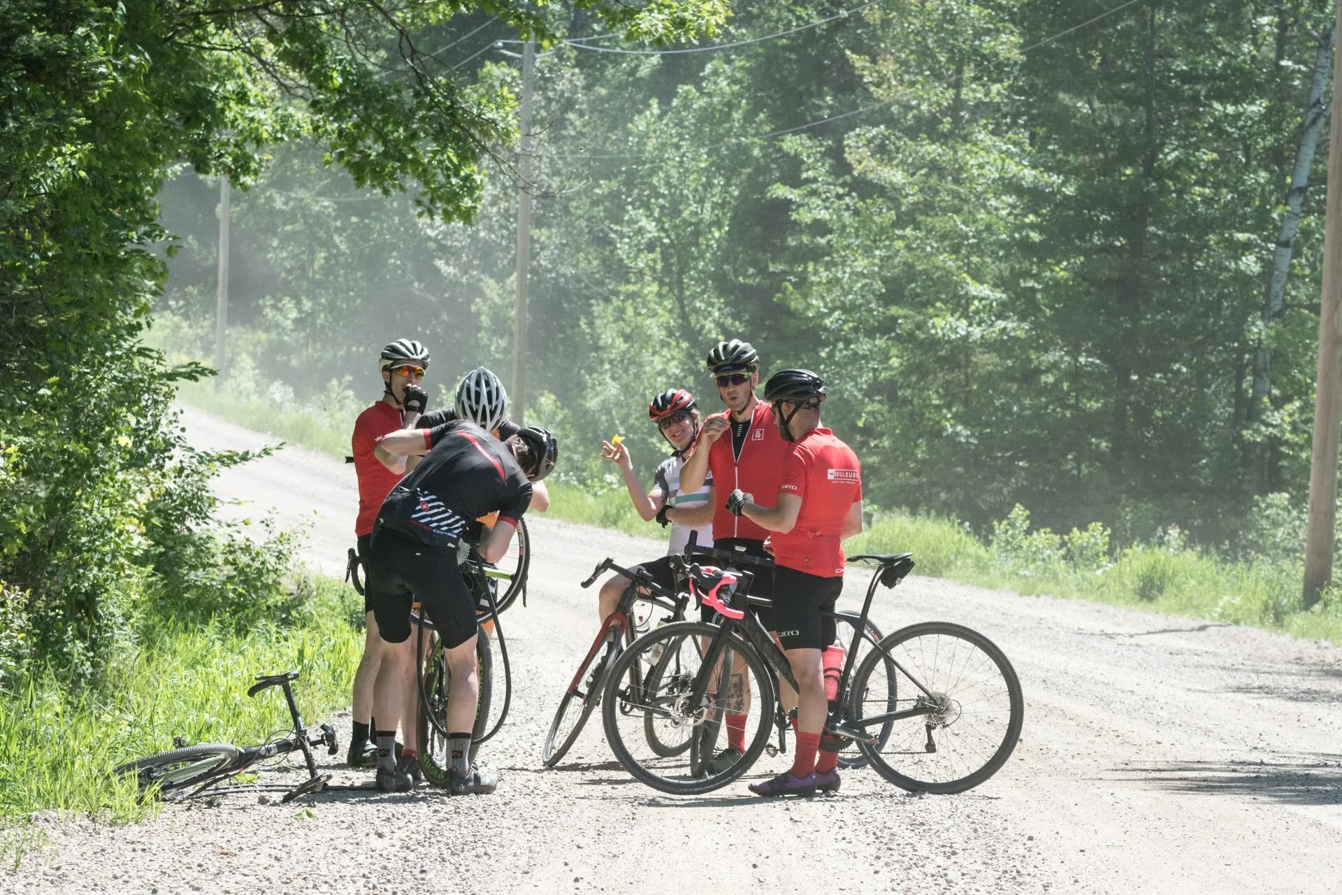 Group of five cyclists taking a break on a gravel road in a wooded area, with fallen bike on the ground.
