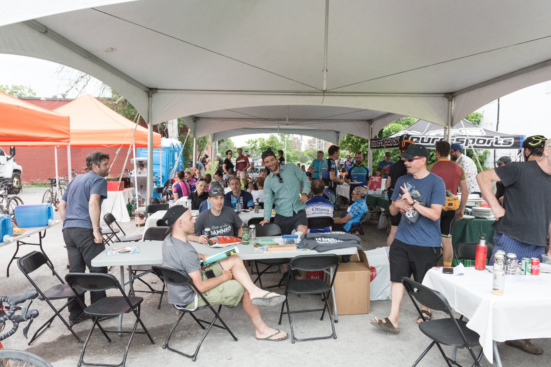 People gathering under a large tent at an outdoor event, with some seated at tables, and others standing and talking, in front of bicycles and orange tents.