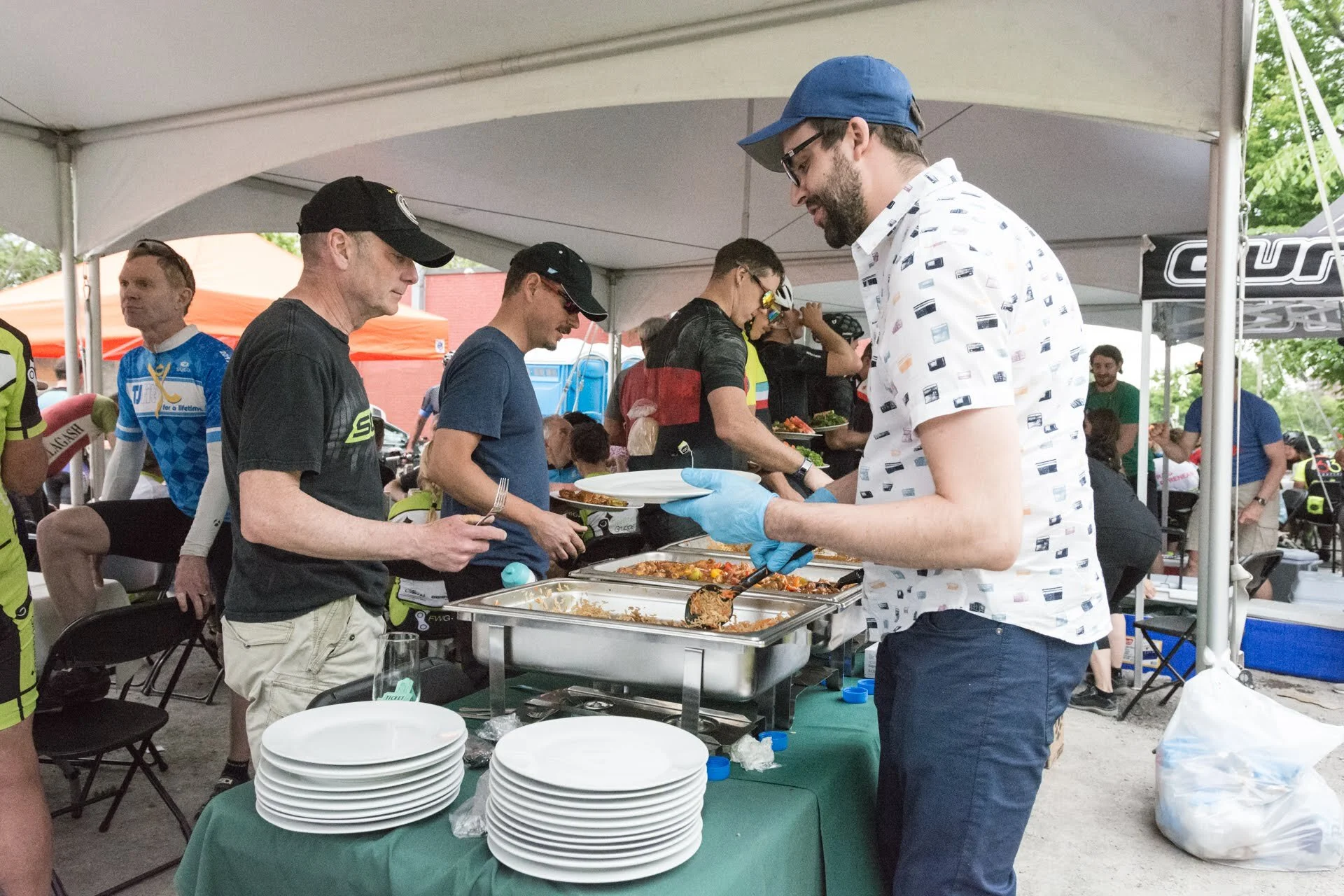People in line receiving food at a buffet table under a tent, with servers serving dishes. There are orange and white tents in the background and some people are seated at tables.
