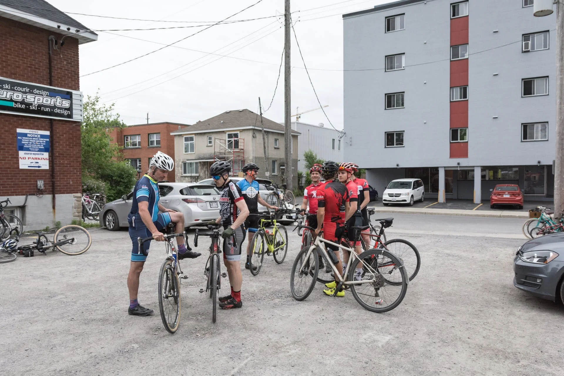 Group of cyclists in colorful jerseys and helmets standing with their bikes in a parking lot, with residential buildings and parked cars in the background.