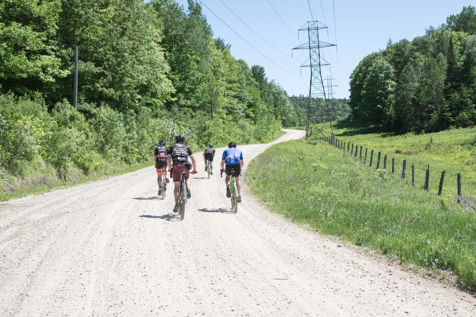 Four people cycling on a dirt road surrounded by green trees and grass, with power lines in the background on a sunny day.