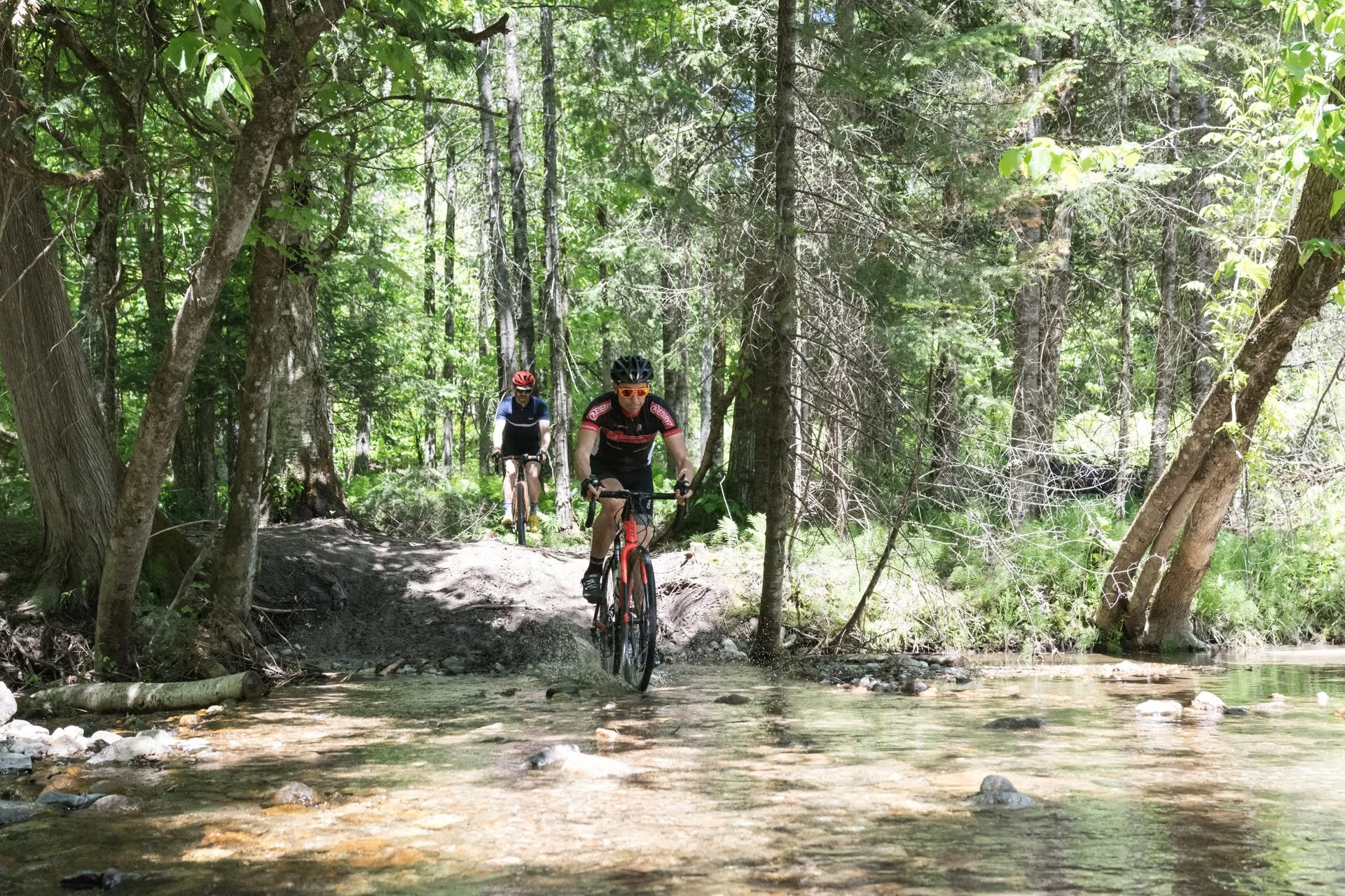 Two cyclists riding mountain bikes through a forested trail crossing a shallow creek.