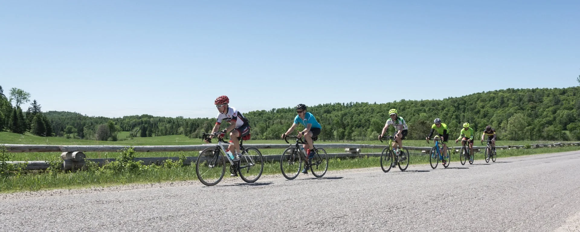 Group of six cyclists riding on a rural road with green trees and hills in the background on a sunny day.