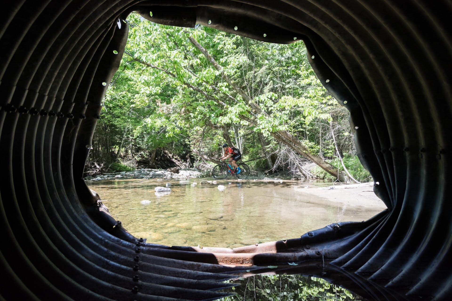 View of a cyclist crossing a shallow creek, seen through a corrugated metal pipe, with lush green trees in the background.