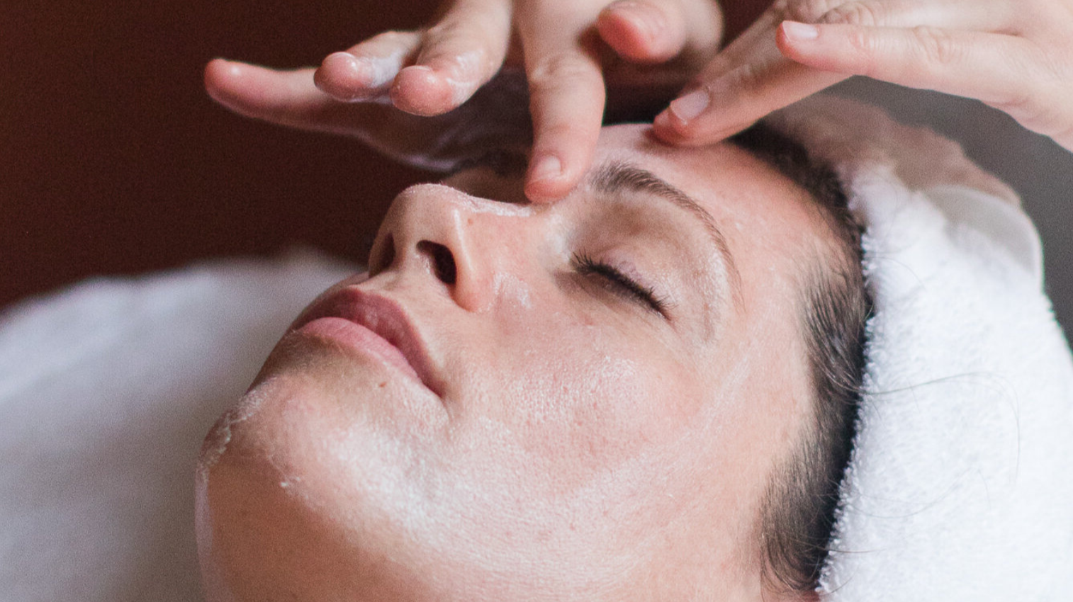 A woman receives a facial treatment at a spa, lying with eyes closed while a skincare professional applies a product on her face.