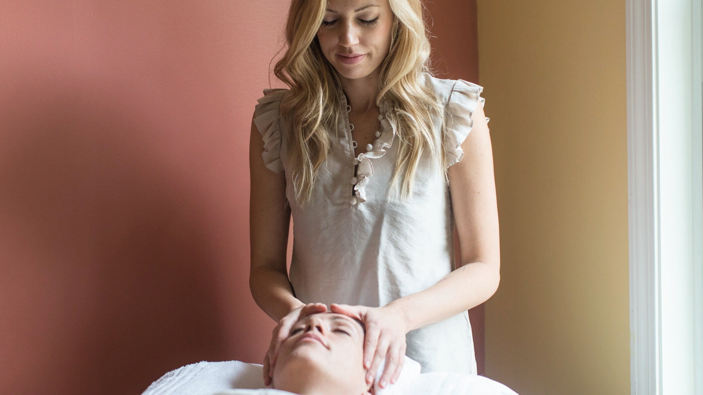 A woman giving a facial massage to a person lying down with their eyes closed, in a room with warm-colored walls and natural light from a window.