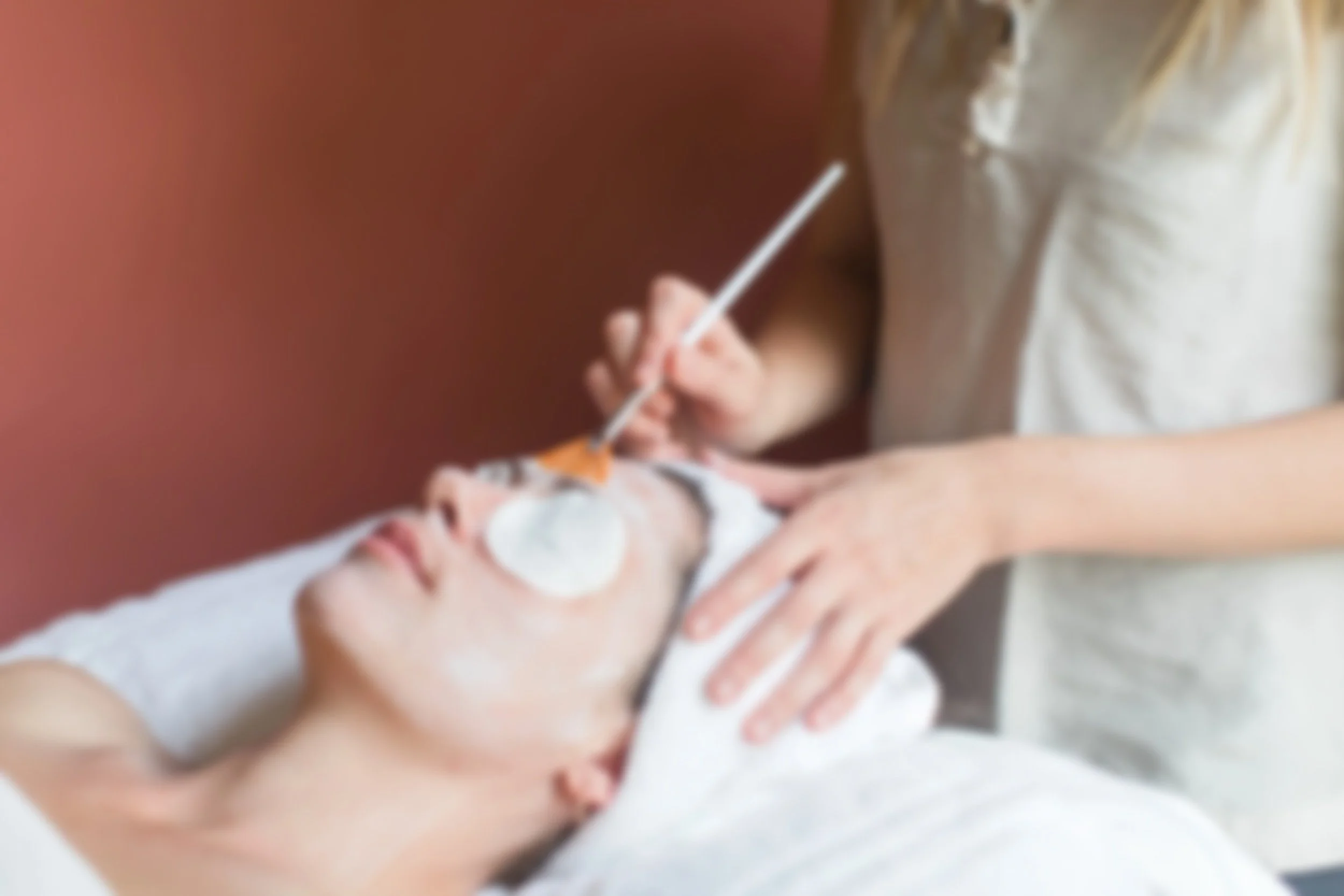 A woman receives a facial treatment with cotton pads on her face while a practitioner applies a mask or product using a brush in a spa or skincare clinic.