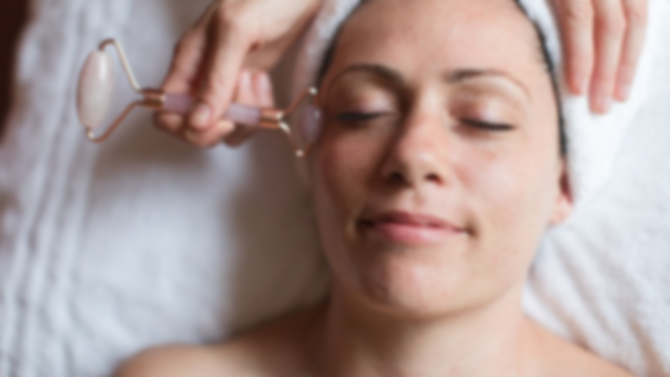 Woman receiving facial massage with jade roller, relaxed and smiling with closed eyes, lying on a bed with a white towel and headband.