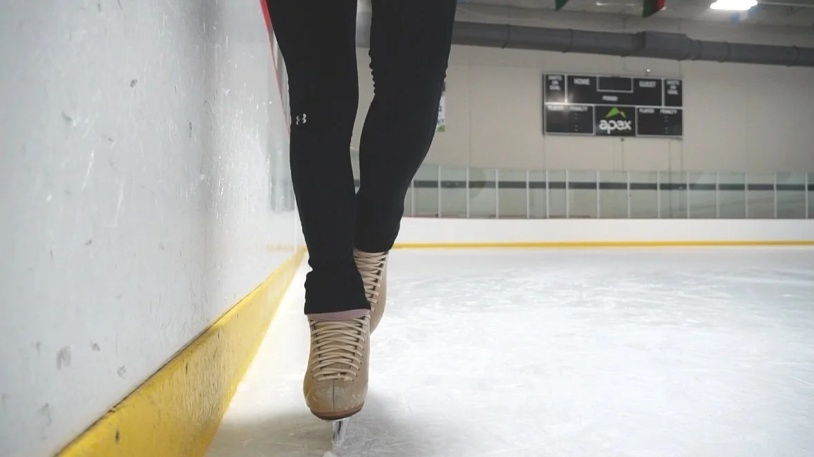 Close-up of a person in black leggings and beige ice skates skating on an indoor ice rink with a black scoreboard in the background.
