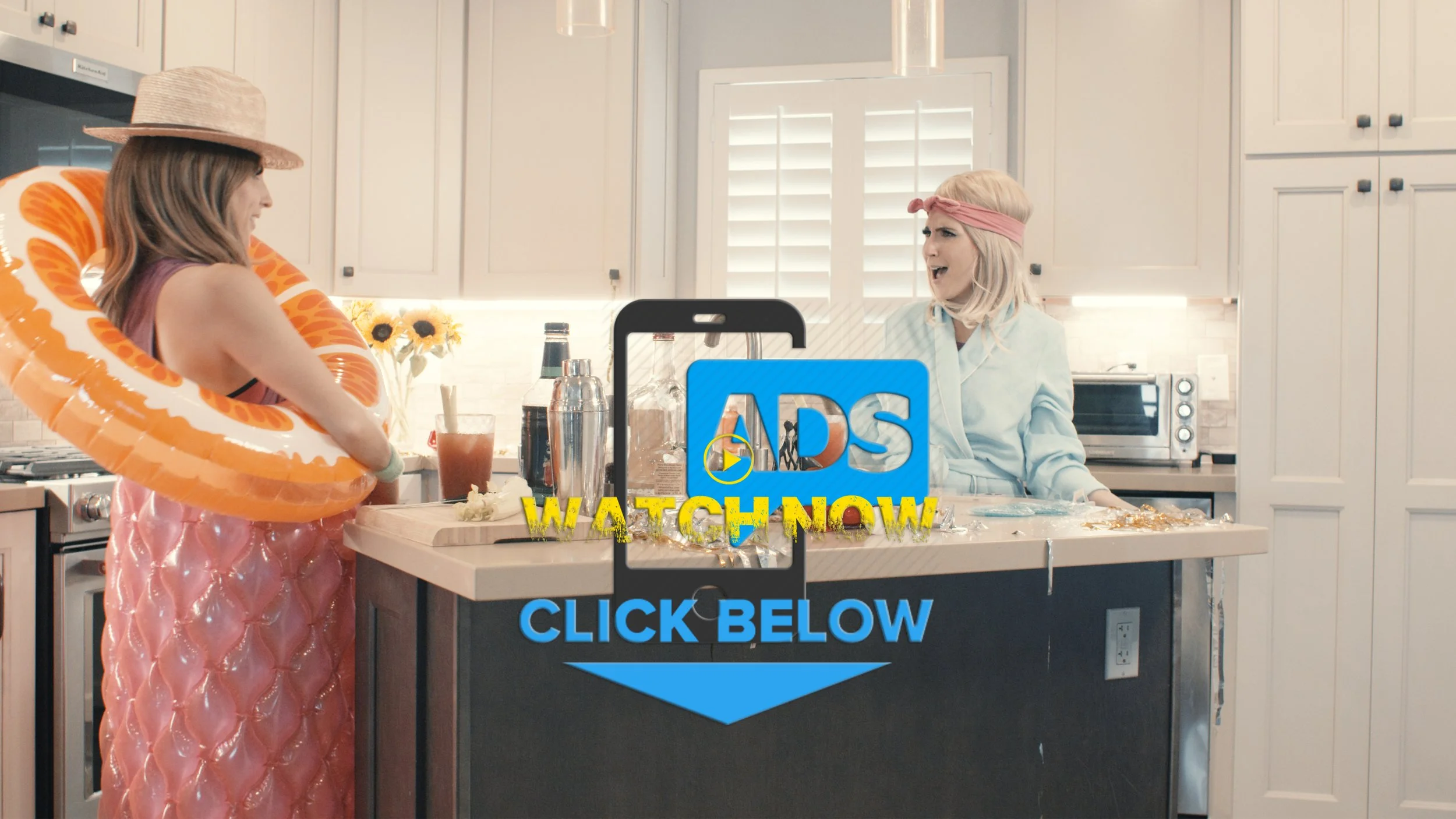 Two women dressed in costumes are in a kitchen, one wearing a pink dress with a large inflatable orange and white float around her waist, and the other wearing a light blue outfit with a pink headband, appearing distressed. The kitchen is modern with