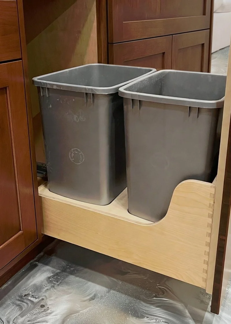 Two gray trash cans placed on a wooden pull-out shelf in a kitchen cabinet.
