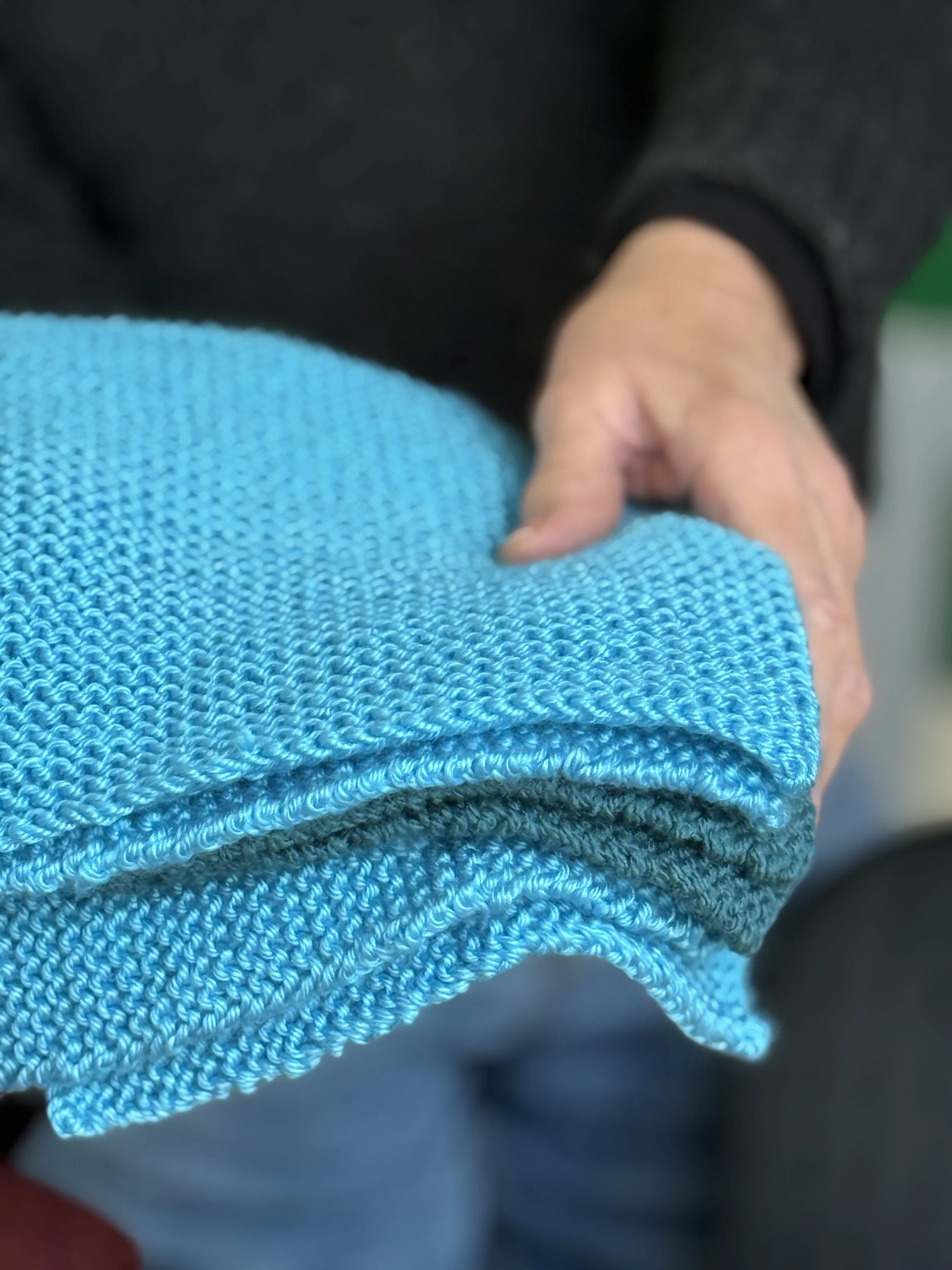 Close-up of a person's hand resting on their lap, with a blue textured fabric of clothing visible in the foreground, and a darker garment on the person's arm.