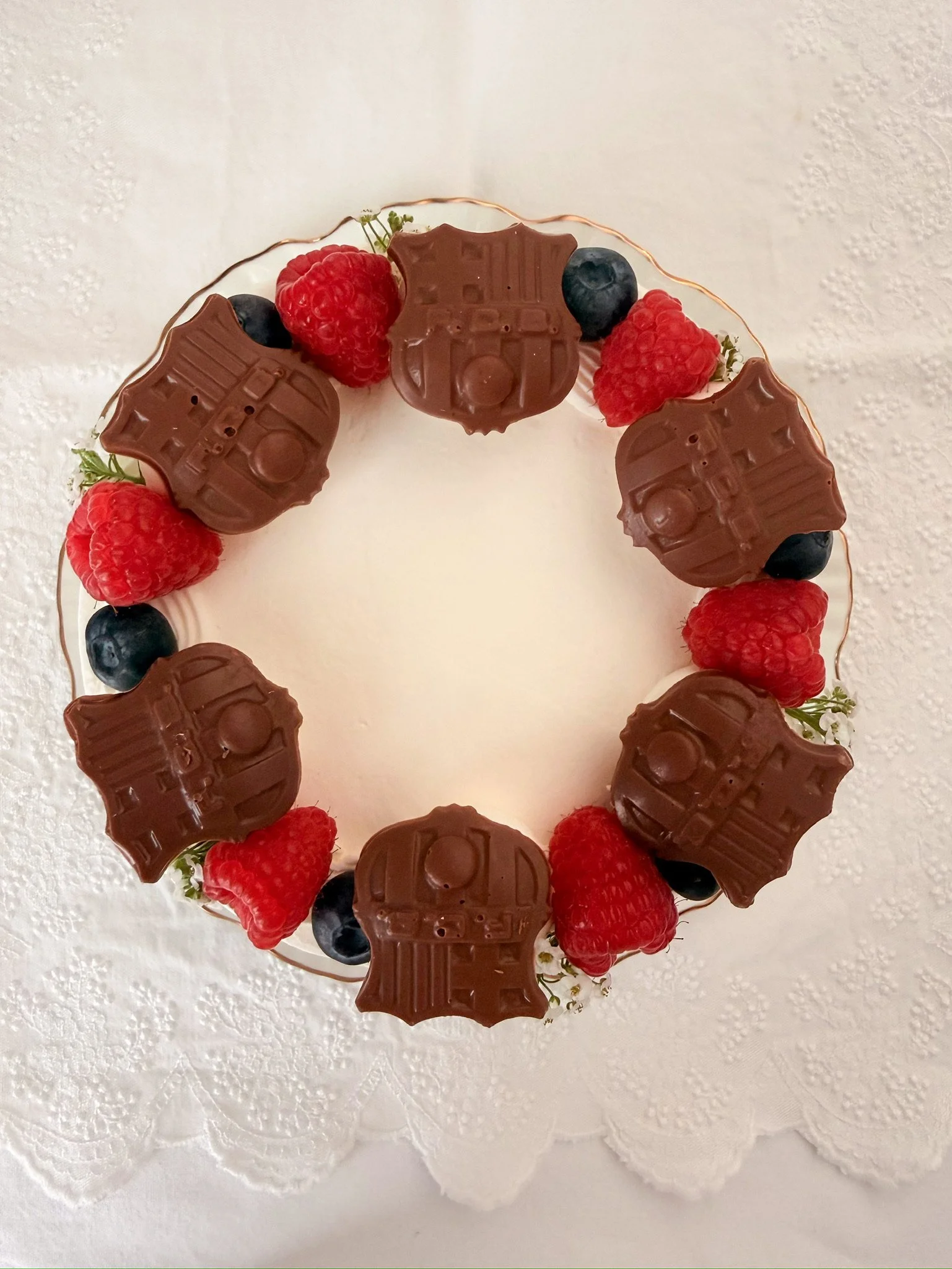 Decorated white cake with raspberries, rose petals, and cream dollops on a gold-rimmed white plate.