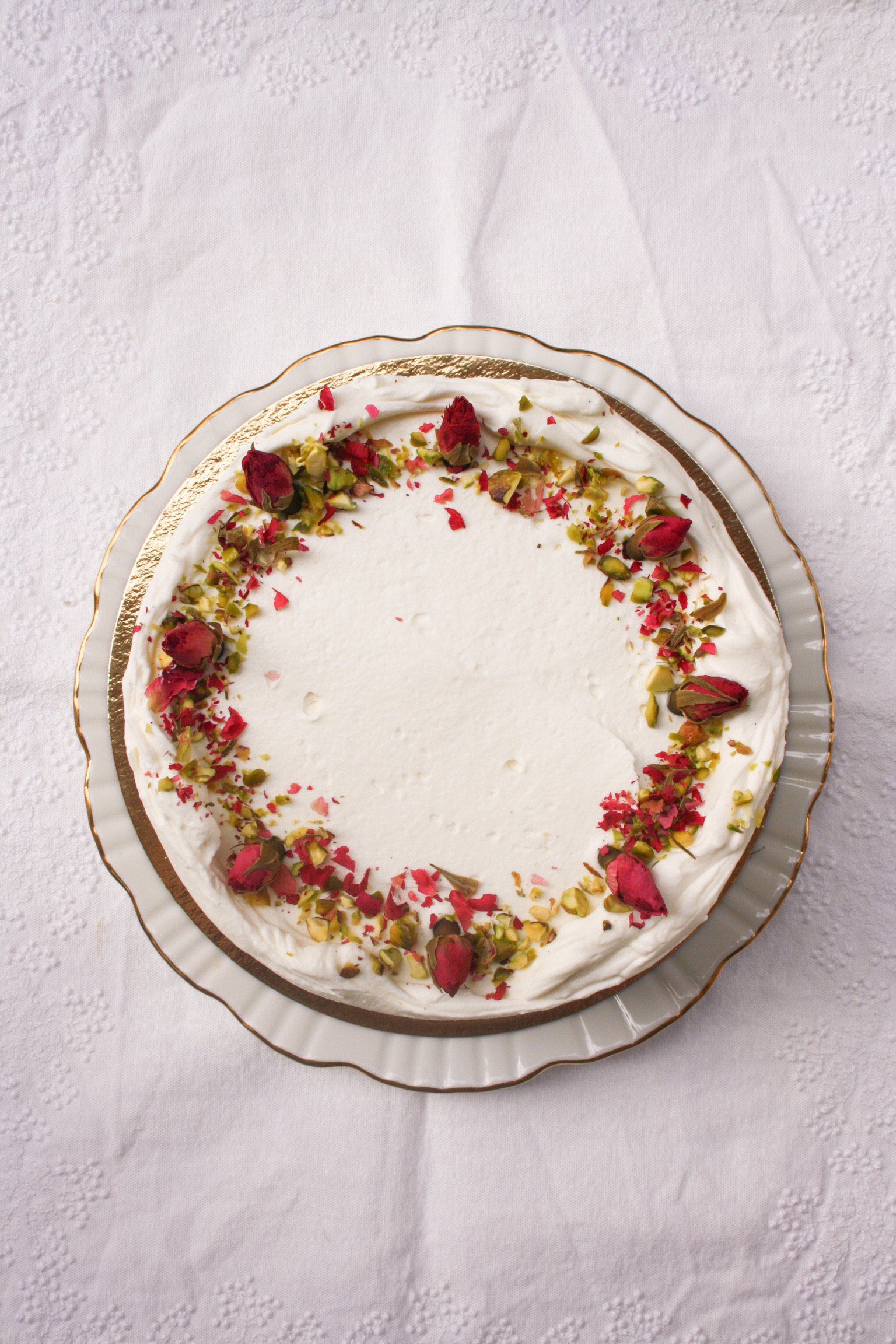 Top view of a decorated cake with white frosting, pink rosebuds, and chopped pistachios on a white plate with gold trim on a white tablecloth.