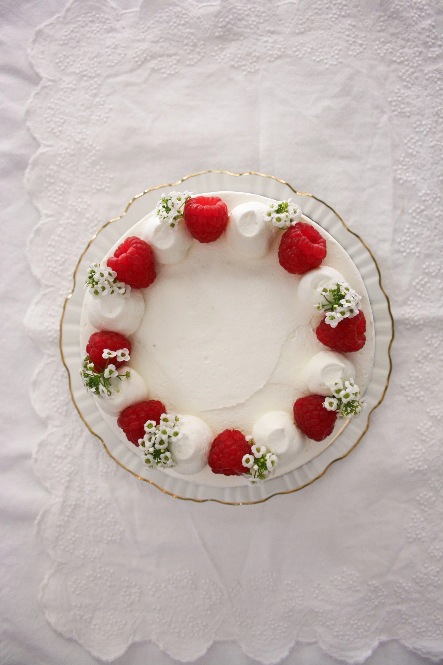 A round white frosted cake decorated with raspberries, small white flowers, and dollops of whipped cream.