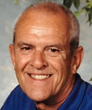 Close-up portrait of a smiling middle-aged man with short gray hair, wearing a blue shirt, against a blurred outdoor background.
