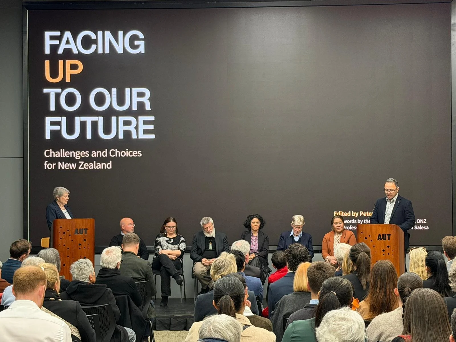 Launch of hte book, Facing Up to Our Future at the AUT Inversity, Helen Clark, and Damon Salesa at podium and 6 authros including Peter Davis sta in between.