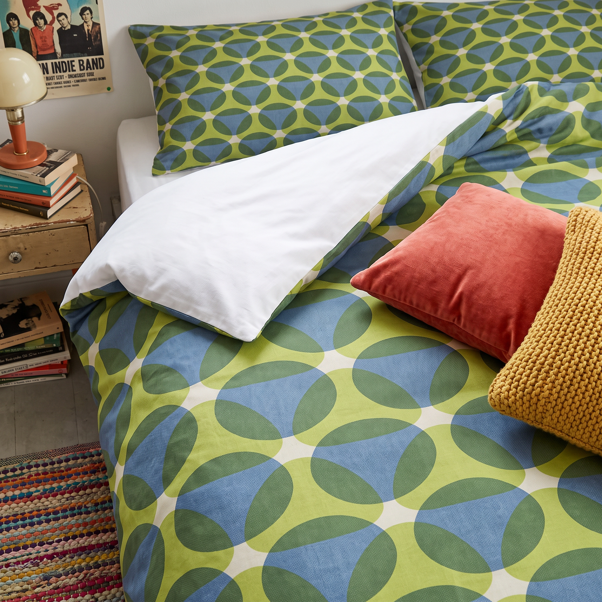 A bed with green and blue patterned pillows and a matching duvet cover, beside a wooden nightstand with books and a vintage lamp. Part of a poster of a band is visible on the wall, and a colorful woven rug is on the floor.