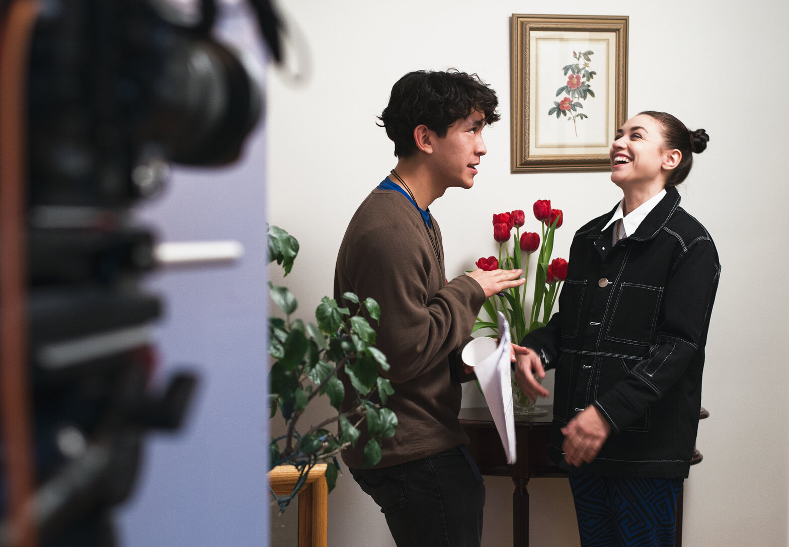A young man and young woman are smiling and talking, with the man touching a bouquet of red tulips in a room with framed artwork on the wall.