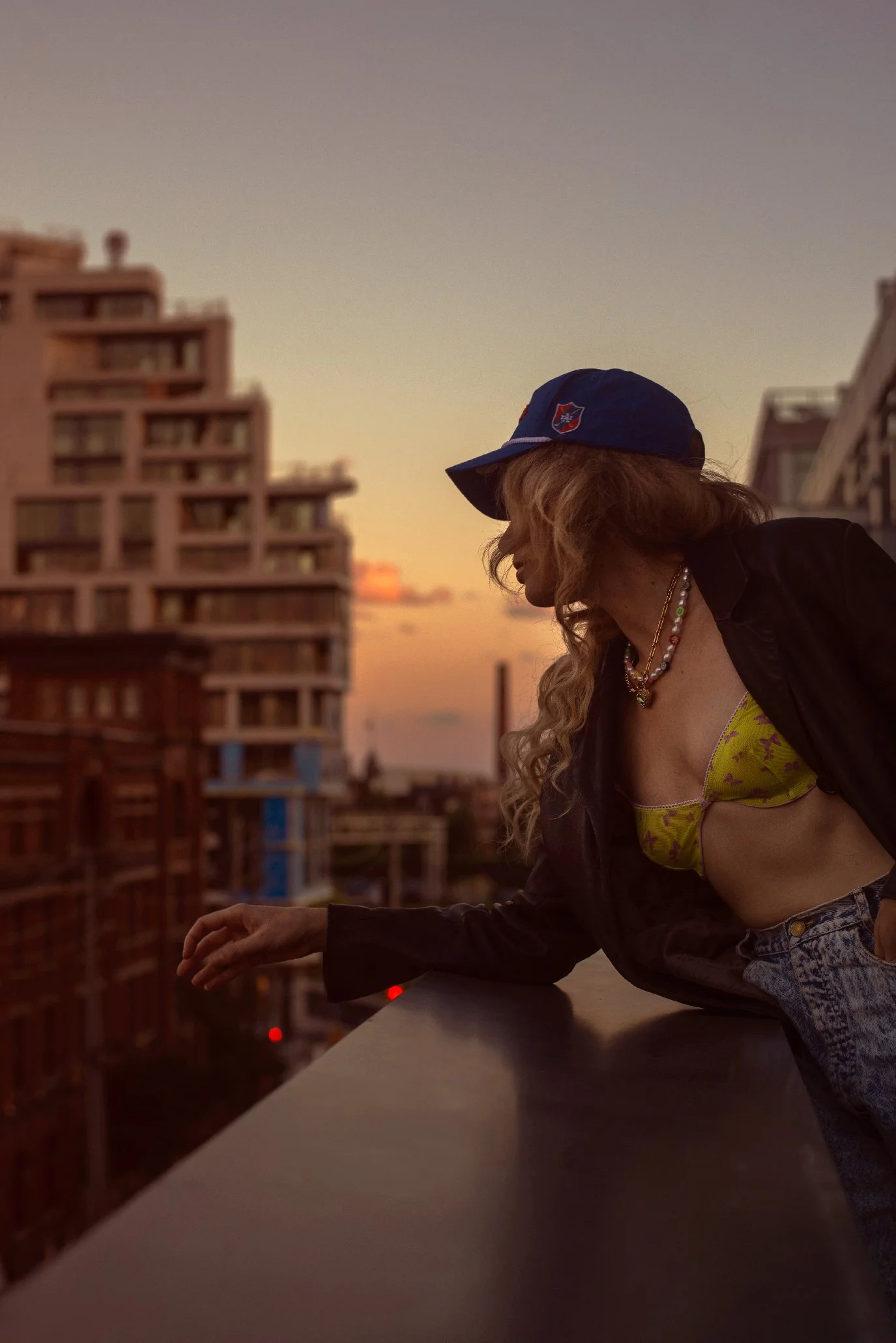 Woman with curly hair wearing a blue baseball cap, black jacket, yellow bra, and colorful necklaces leaning on a balcony railing during sunset in an urban setting.
