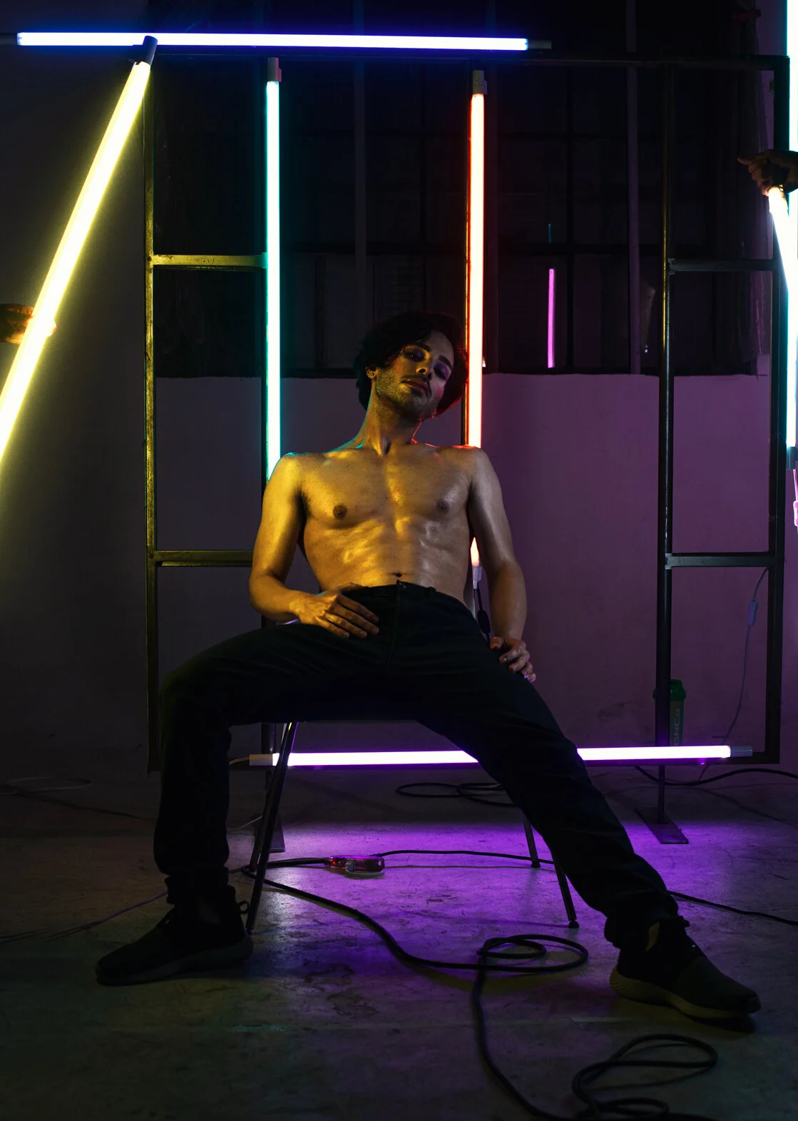 A shirtless man with dark hair and makeup, seated with legs apart and hands on thighs, in front of a neon-lit background with vertical and horizontal light tubes in blue, red, purple, and yellow colors.