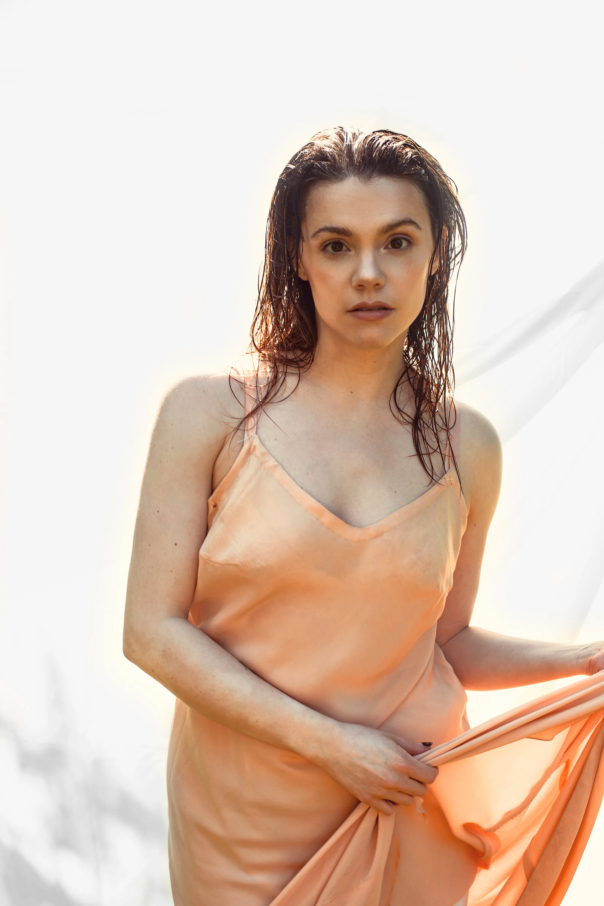 A woman with wet dark hair wearing a peach satin dress, holding the fabric in front of her, standing outdoors with a bright, overexposed background.