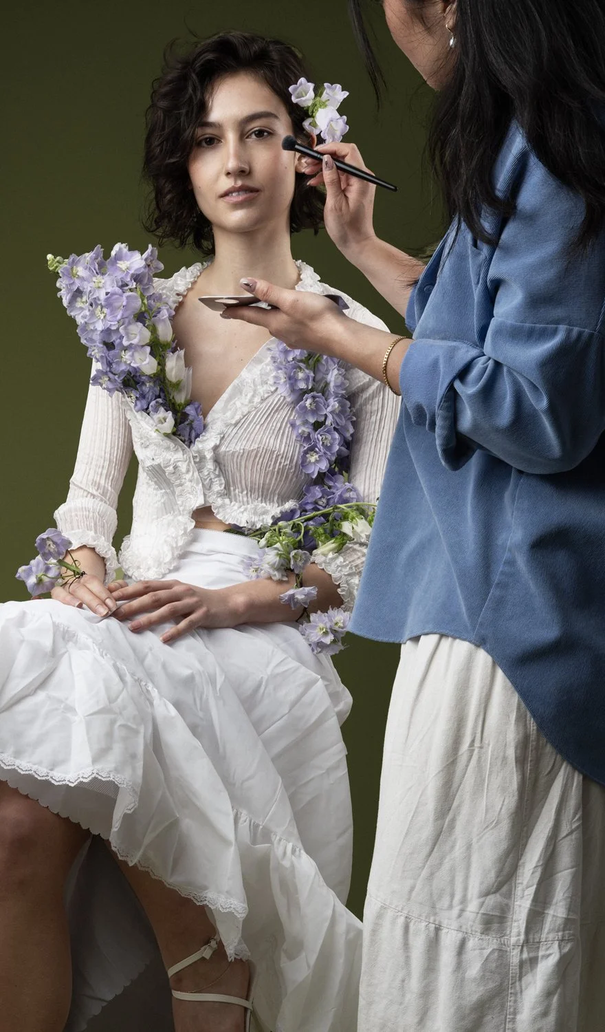 A woman sitting with flowers on her white dress and a makeup artist applying makeup to her face, with a green background. Behind the Scenes