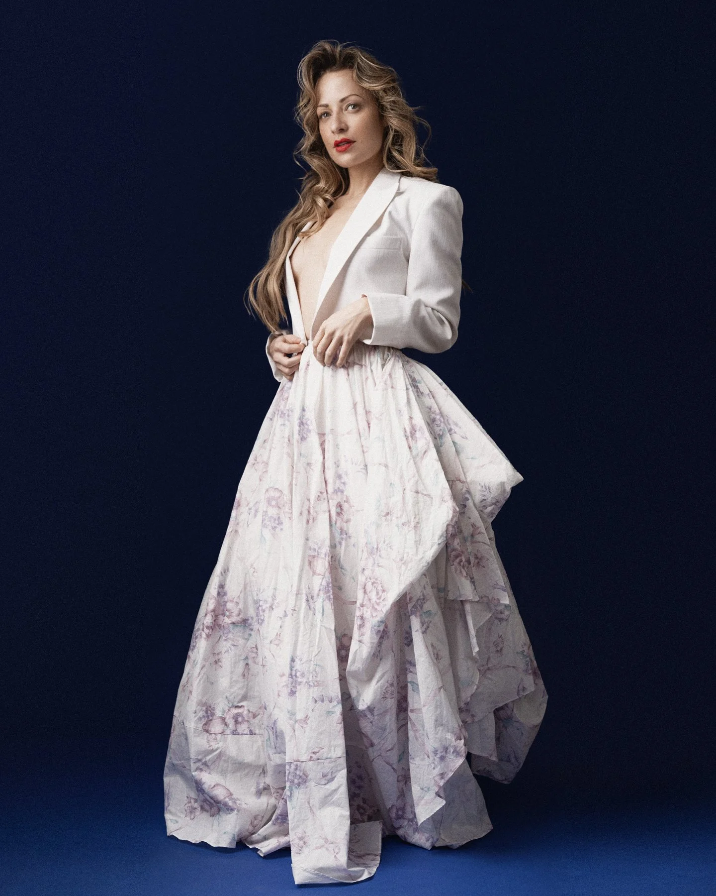 Woman with long wavy hair wearing a white blazer and a floral high-low skirt against a dark background. Karen Knox