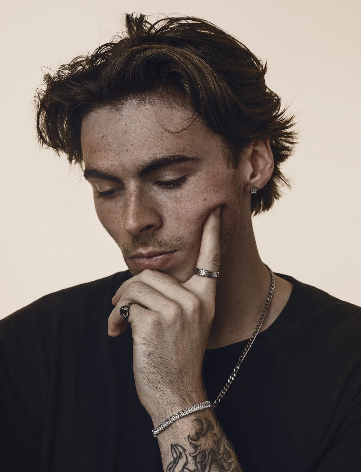 A young man with dark hair, wearing earrings, a chain, and rings, touching his face thoughtfully, with tattoos on his arm. Photo of David July