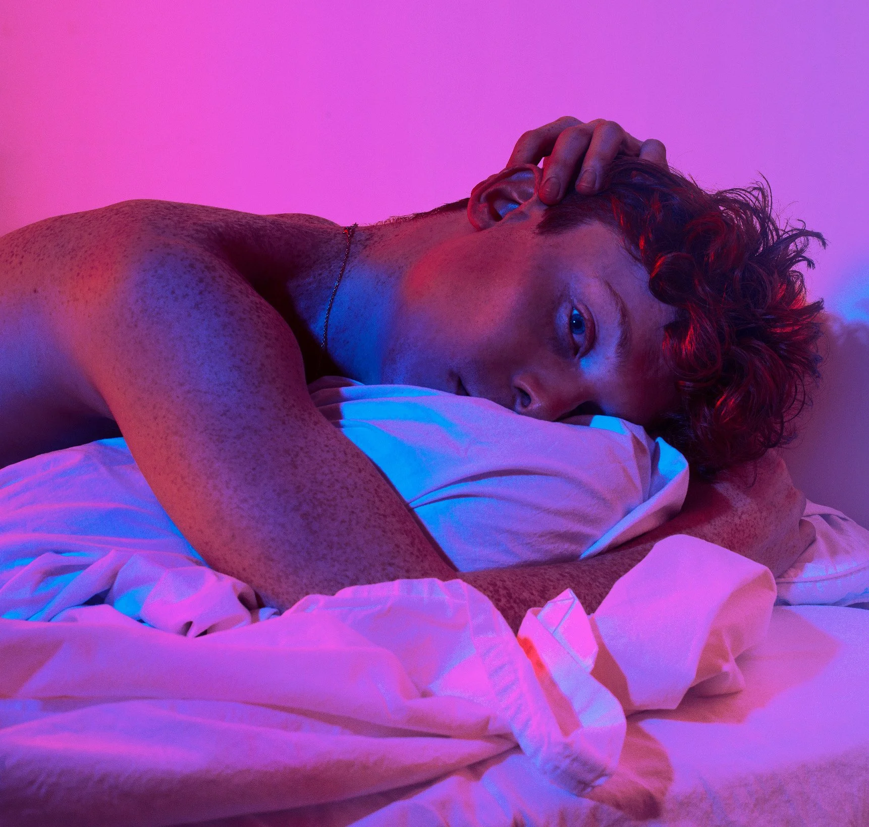 A young man with curly red hair and freckles lying on a bed with pink and blue lighting, resting his head on a pillow, looking into the camera.