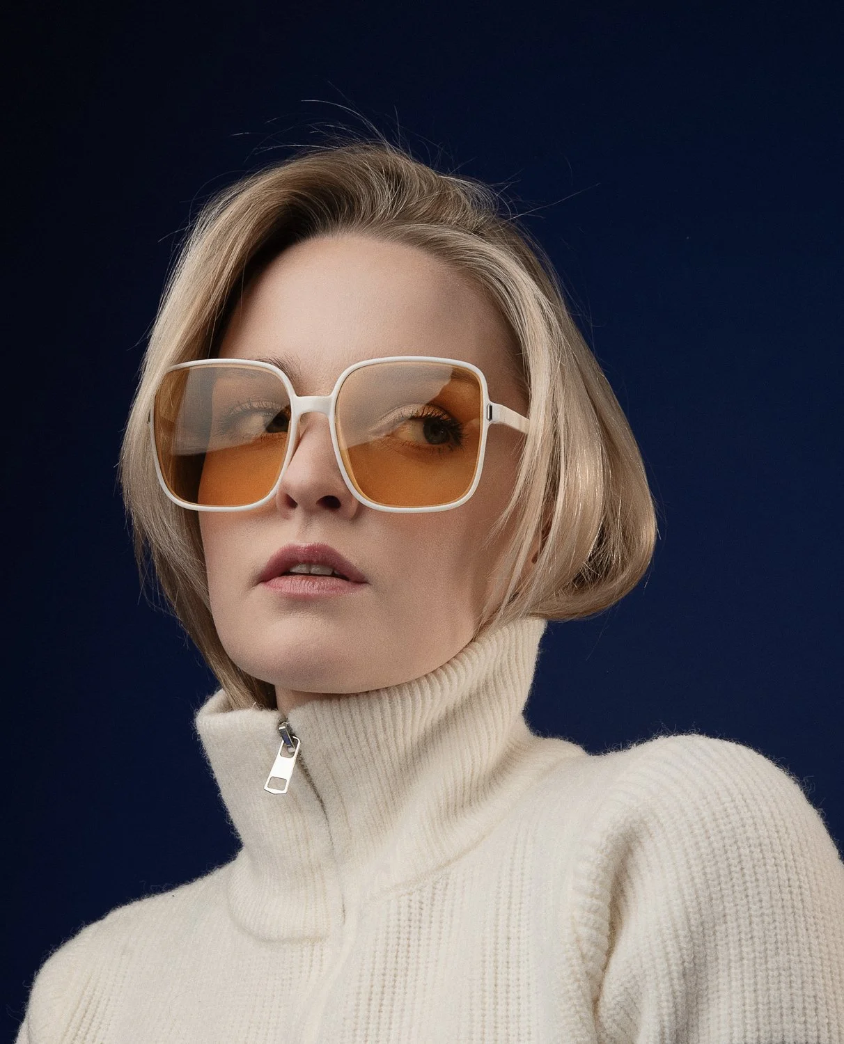 A woman with short blonde hair wearing large rectangular sunglasses with orange lenses and a cream-colored high-neck sweater against a dark blue background.