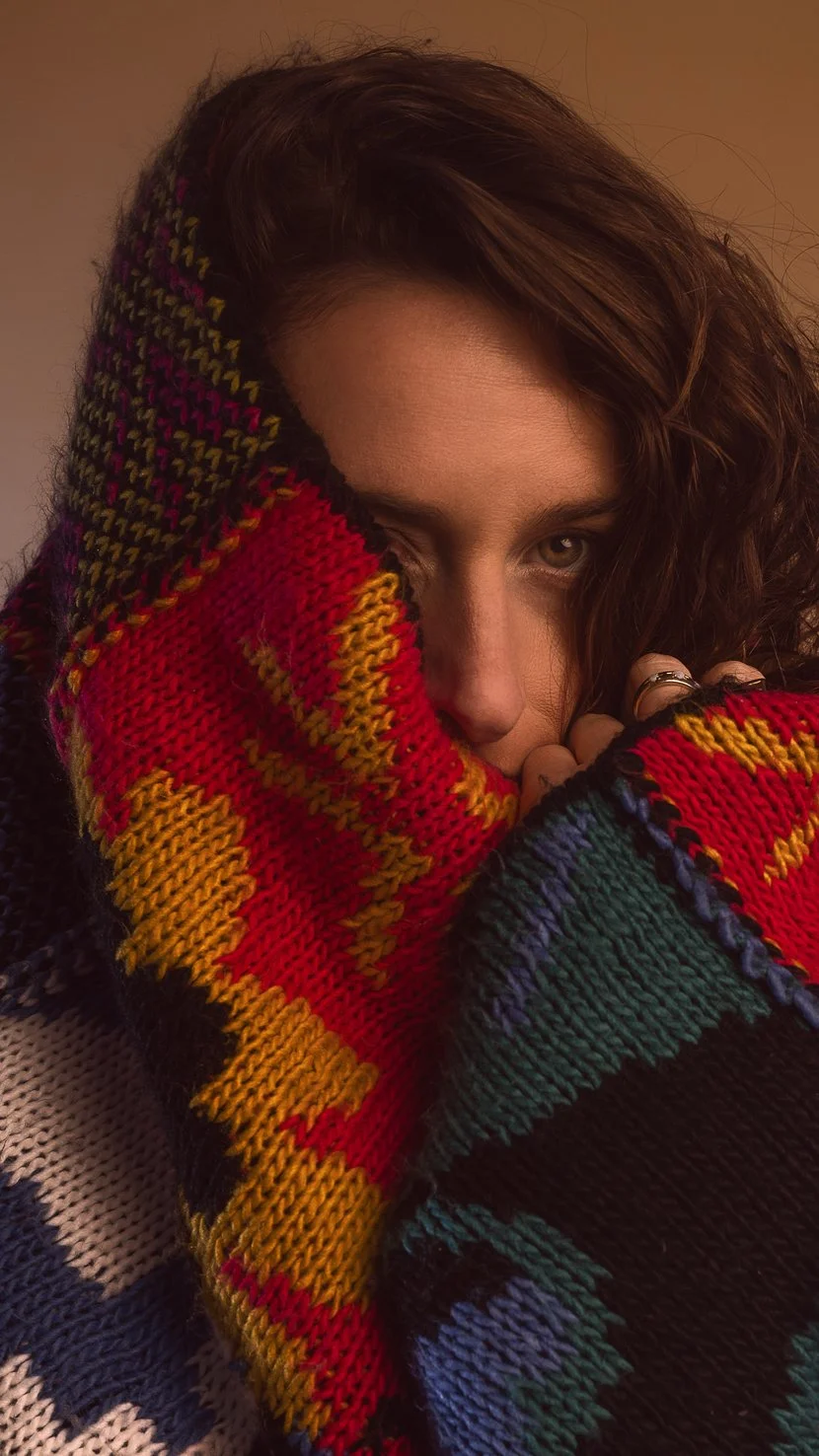 Close-up of a woman with wavy brown hair, partially covering her face with a colorful knitted sweater in red, yellow, green, and blue.