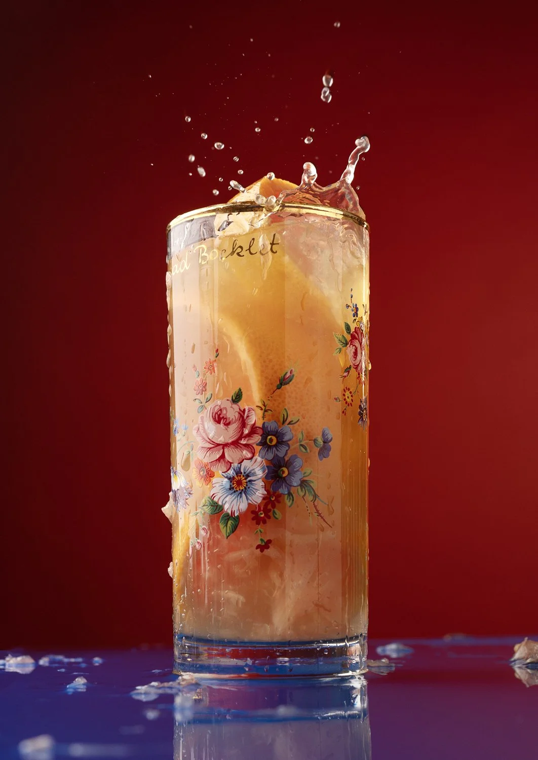 A tall glass with a floral pattern contains a beverage with lemon slices, ice, and floating fruit pieces, with water splashing at the top against a red background.