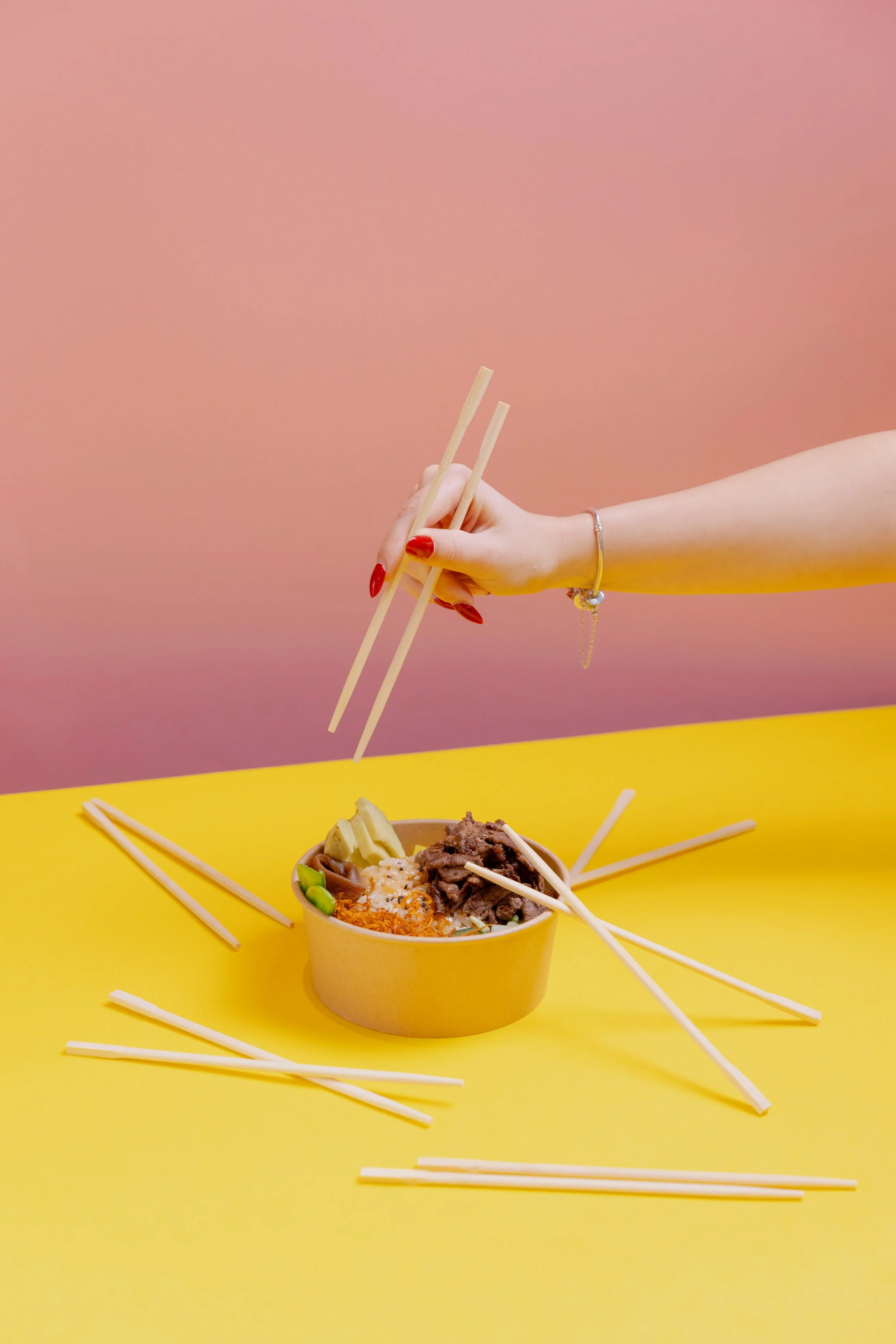 A person's hand holding chopsticks above a bowl of ramen, with several chopsticks scattered on a yellow surface, pink background, and wearing a bracelet. Ichigo Budapest, Gasztro fotó, marketing fotó, socialmédia, instagram