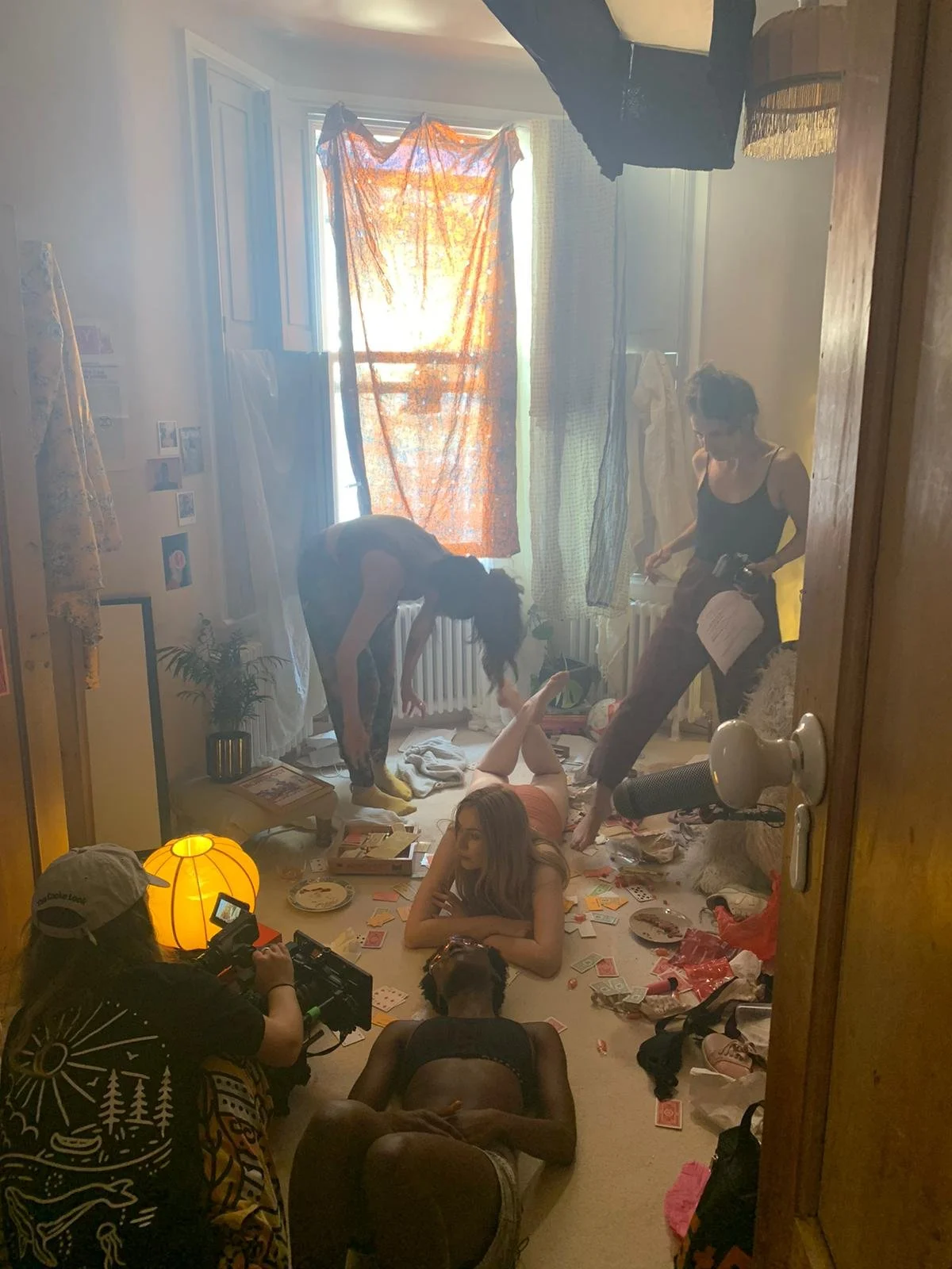 A room filled with people, including a woman lying on the floor, two women standing over her, a person filming, and another person lying on the floor, with laundry hanging near a window, clutter scattered on the floor, and a yellow lamp in the corner